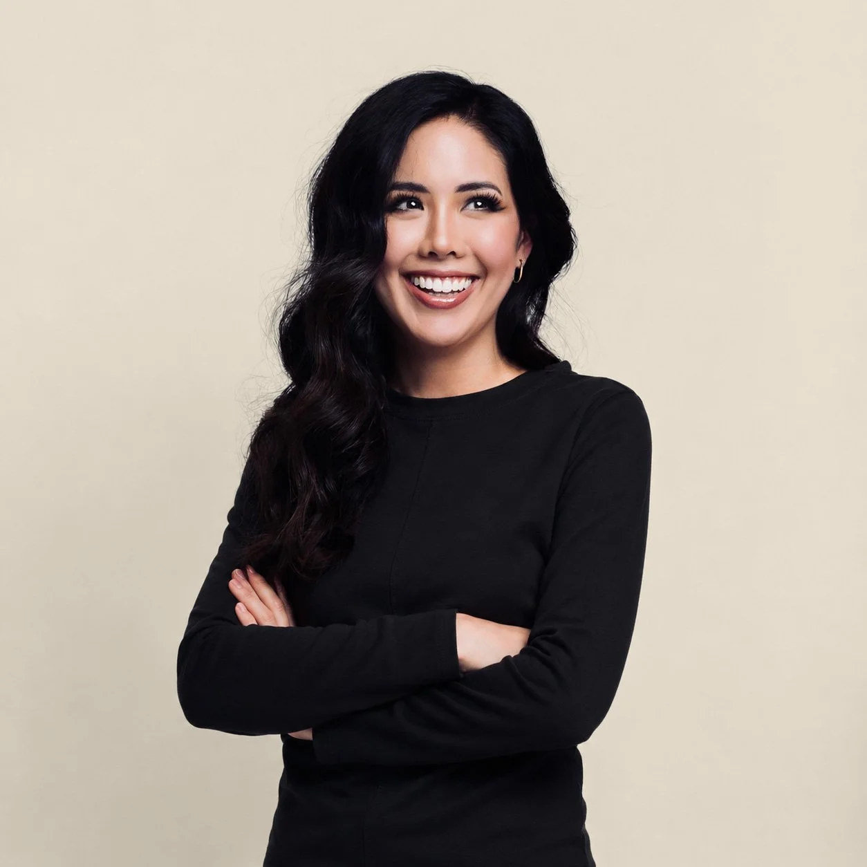 A smiling woman with long dark wavy hair wearing a black long-sleeve shirt, standing against a plain light background.