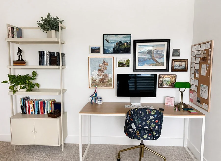 Home office setup with a desk, computer monitor, bookshelf with books and plants, picture frames on wall, and a bulletin board.
