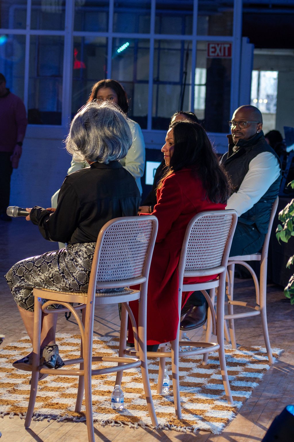 Group of five people sitting on chairs in a discussion or panel in a lit indoor space with large windows and an 'EXIT' sign visible in the background.