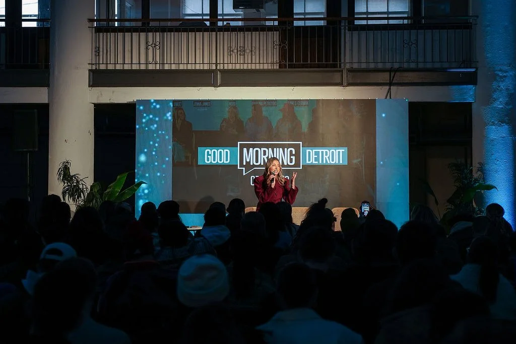 A woman speaking on stage at an event with a large screen behind her displaying the text "Good Morning Detroit." The audience is seated in front of her, and there are two plants on either side of the stage.