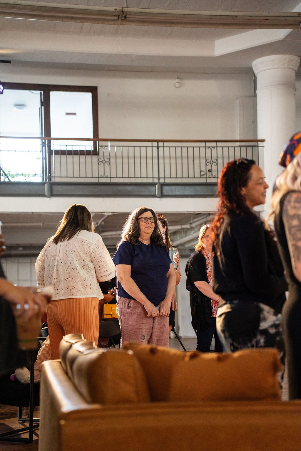 Group of women standing and talking inside a well-lit room with a high ceiling, metal railing, and large window in the background.