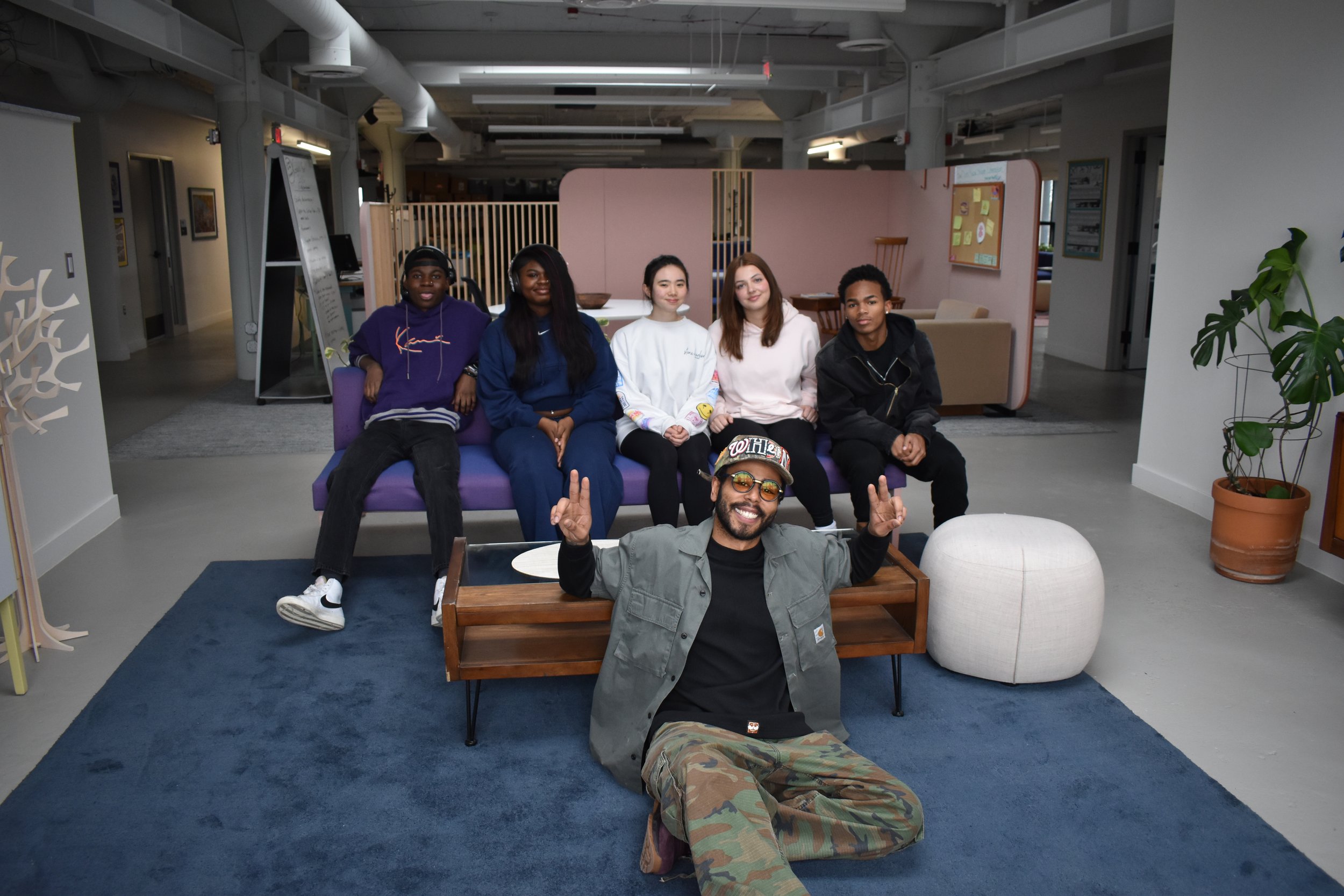 A group of six diverse young people, five women and one man, posing in an indoor lounge area with a purple sofa, a wooden coffee table, and a large potted plant. They are sitting and lying on the furniture, smiling and enjoying their time together.