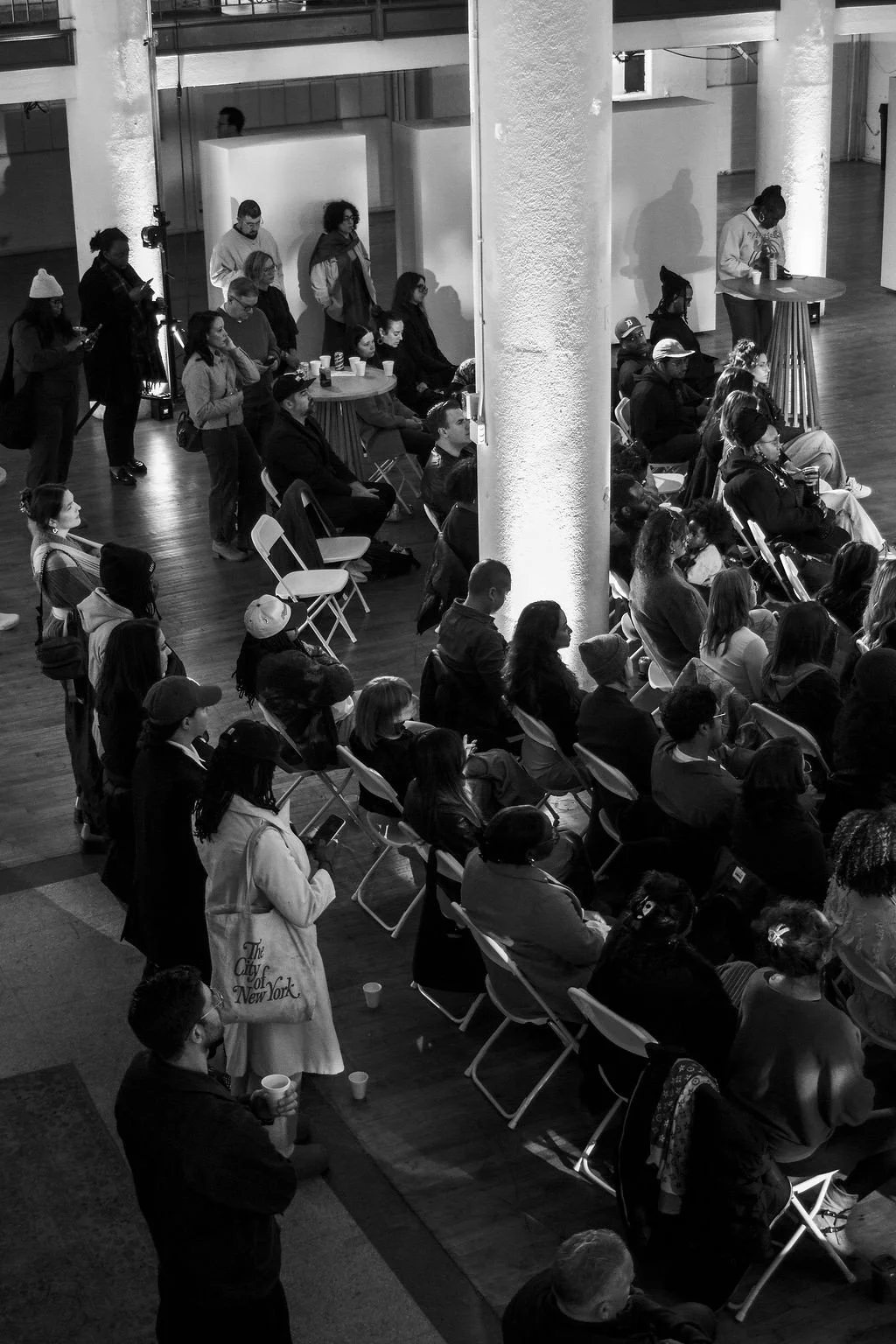 Black and white photo of audience members seated and standing, attentively watching an event in a spacious indoor venue with large white columns.