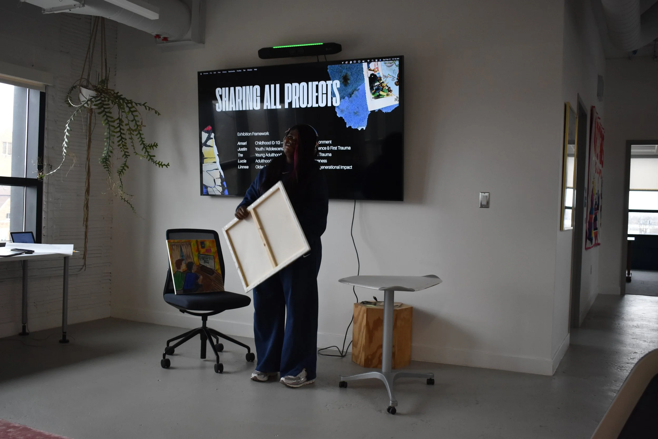 A woman in blue sweats holding a white art frame stands in front of a large screen displaying a presentation titled 'Sharing All Projects.' There is a chair with a painting leaning on it, a table with a laptop, and some paintings on the walls in a well-lit room.