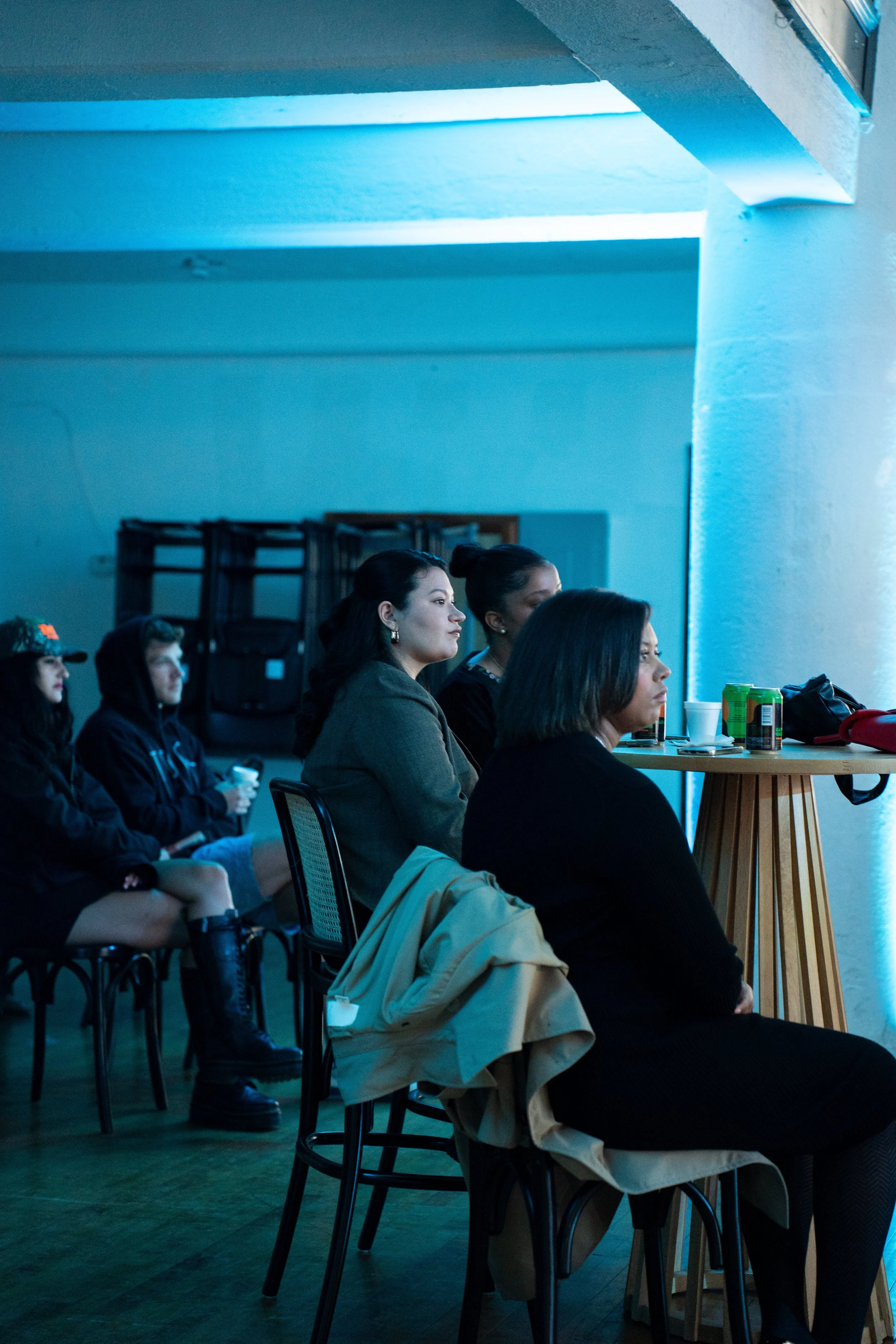 People sitting in a dimly lit room, facing forward, attentively listening to something. Several are holding cups, with cans and cups on a table to their right. The room has a blue hue and looks like a seminar or presentation setting.