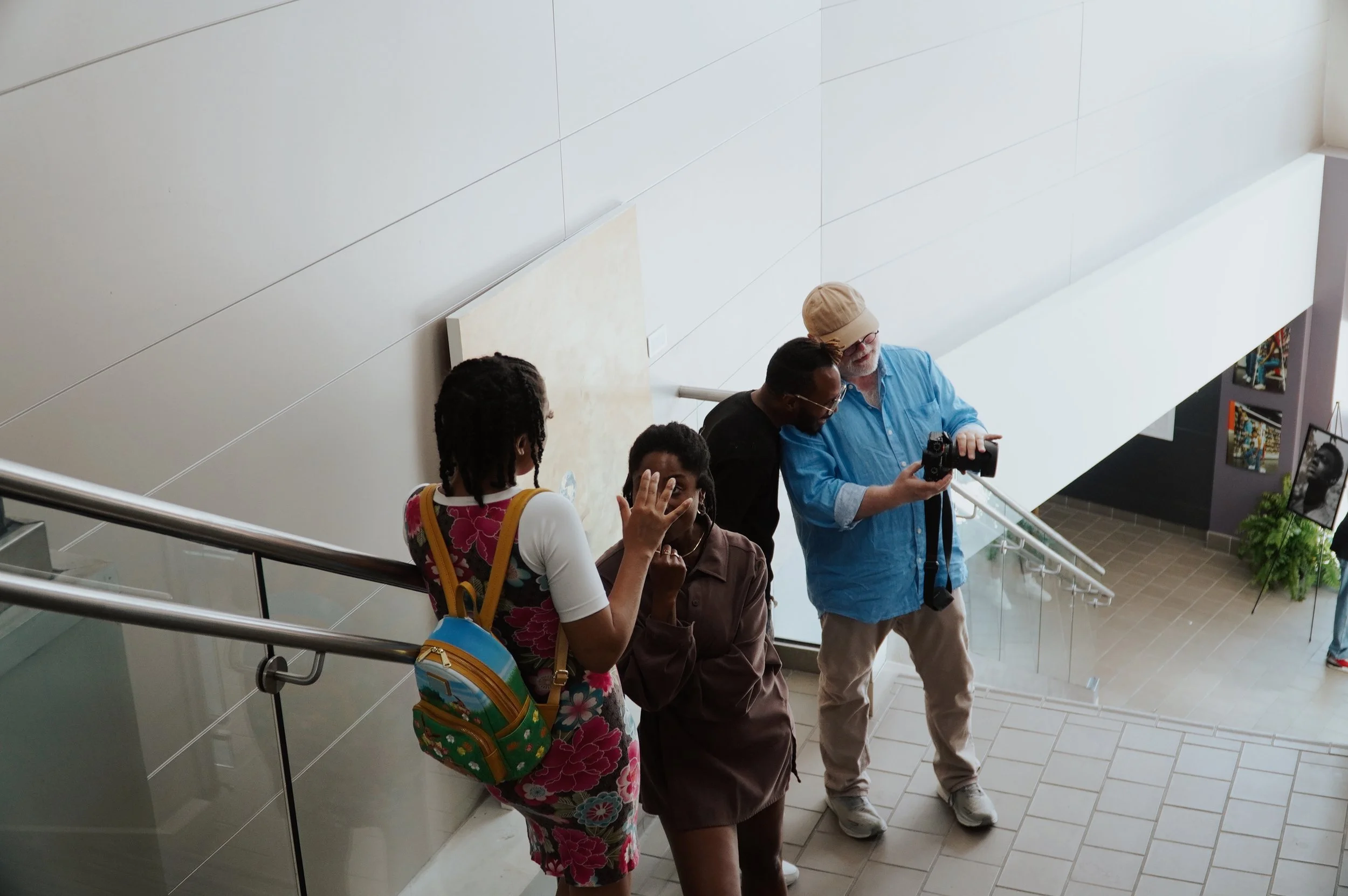 Four people standing in an indoor space near a staircase railing, with a plain wall on one side and a display of photographs on the other. They are engaged in conversation, with some looking at a camera. The group includes a woman with a backpack, a man with glasses and a camera, a woman with glasses and braids, and an older man with a hat and a beard.