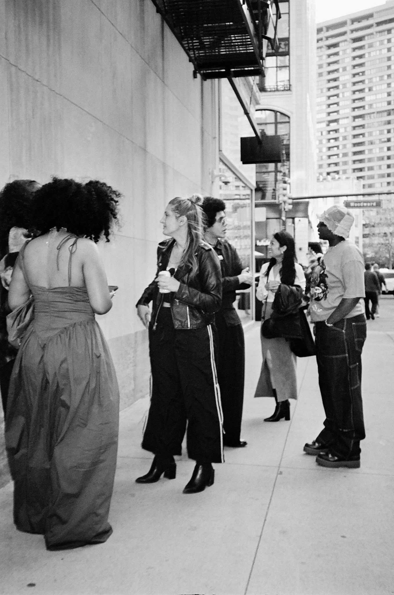 A group of people standing and talking outside a building in an urban setting, with tall buildings and a sidewalk visible in the background. They are wearing fashionable clothing and appear to be engaged in conversation.