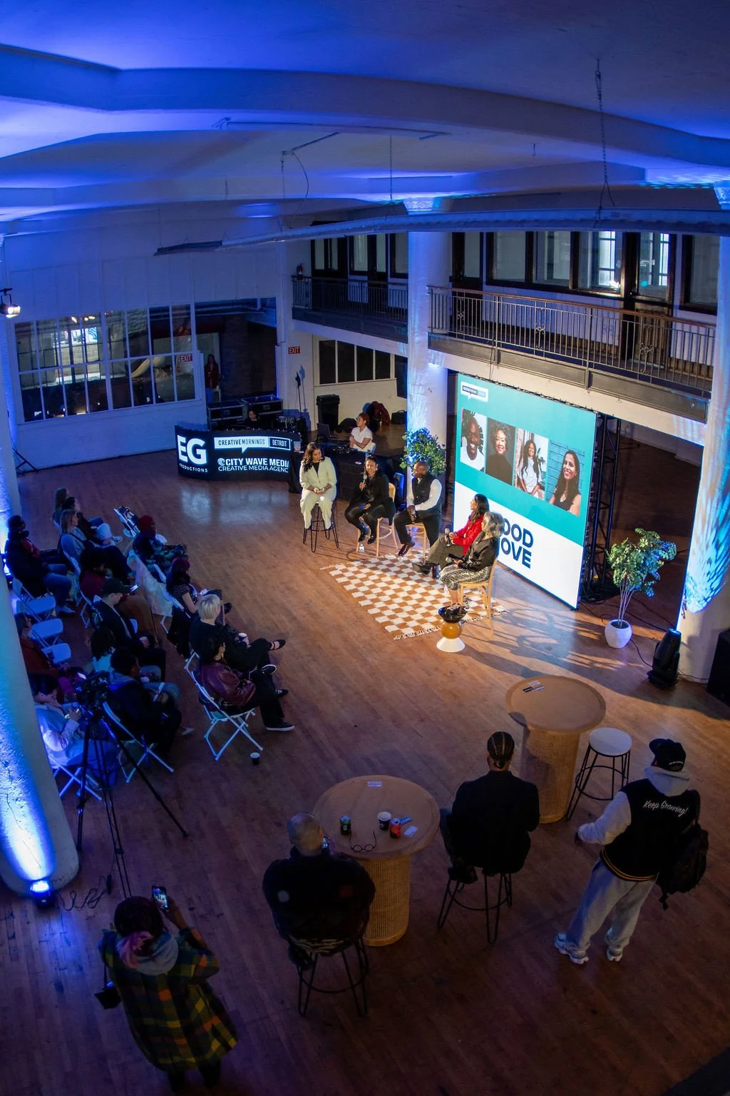 An indoor event with a panel discussion in a well-lit, modern venue. Panelists are seated on the stage, and a large screen behind them displays images and text. Audience members are seated facing the stage, and some are standing or taking photos. The space has wooden flooring, potted plants, and blue accent lighting.