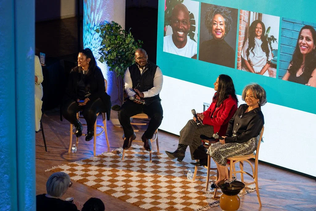 Four people sitting on chairs on a stage, engaged in a panel discussion. Behind them is a large screen displaying photos of five diverse individuals. The stage features a plant and a patterned rug, and there are audience members in the foreground.