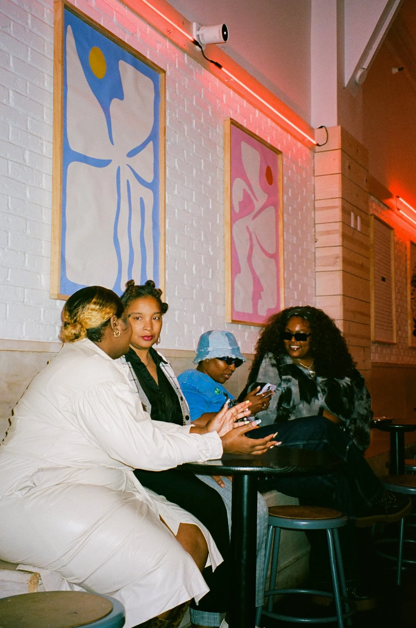 Four women sitting at a bar or cafe, engaged with their phones, with colorful framed artwork on a white brick wall behind them.