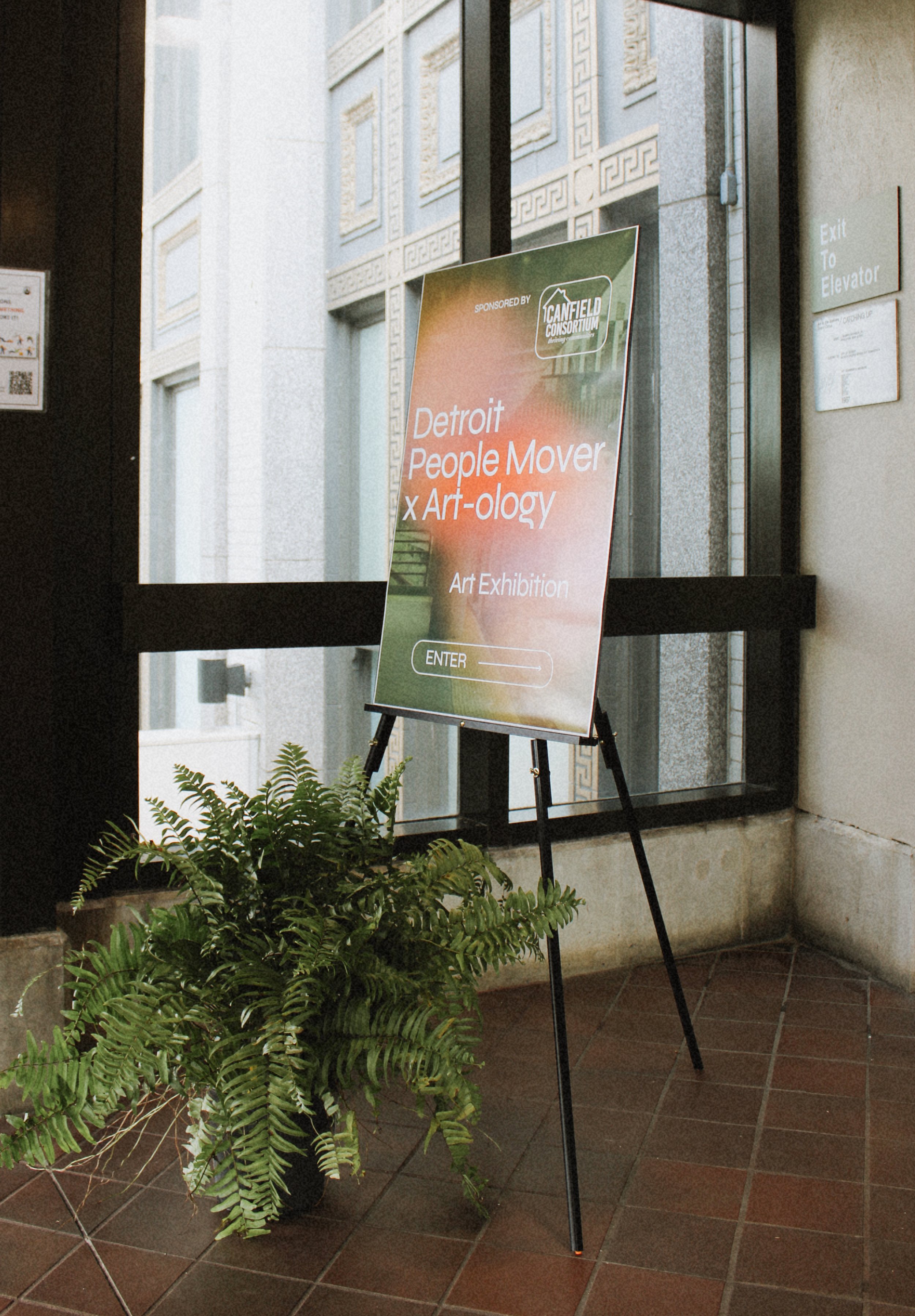 A sign advertising an art exhibition titled 'Detroit People Mover x Art-ology' is displayed on a stand near a potted fern plant inside a building. The background includes a glass door with an 'Exit to Elevator' sign.