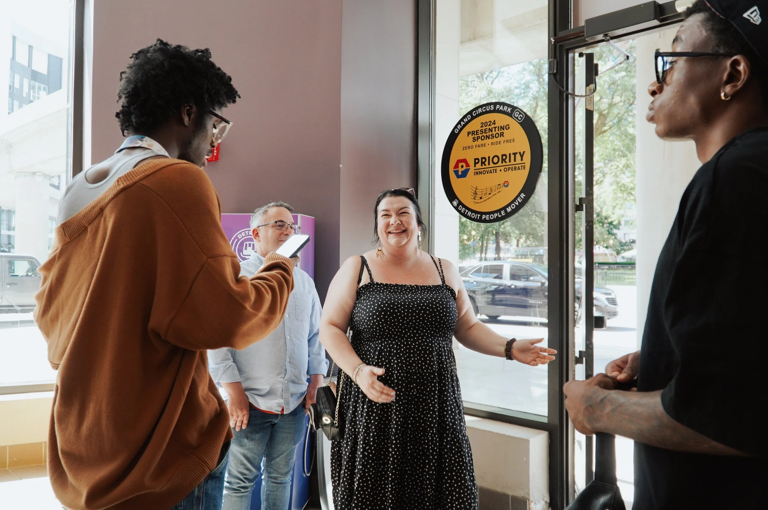 Four people standing by the entrance of a building, engaged in conversation. One woman is smiling and speaking, while others are listening or looking at their phones. The entrance has signs, including a yellow and black one related to the Detroit People Mover.