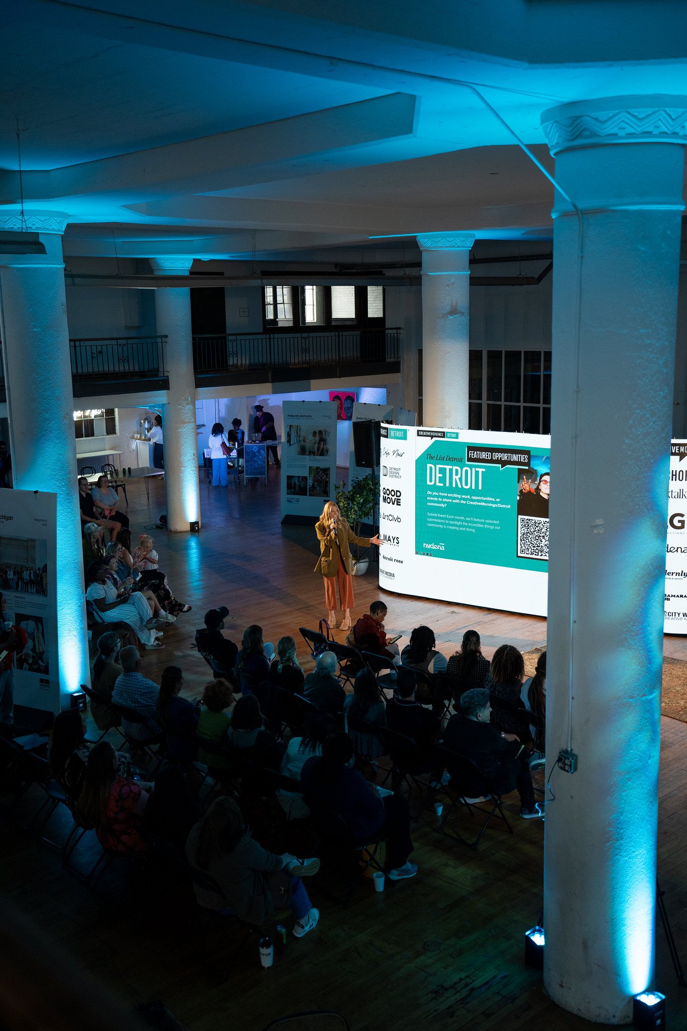 Indoor event space with audience seated and speaker presenting in front of large screen displaying the word 'Detroit'. The room features white columns with decorative capital and blue lighting.