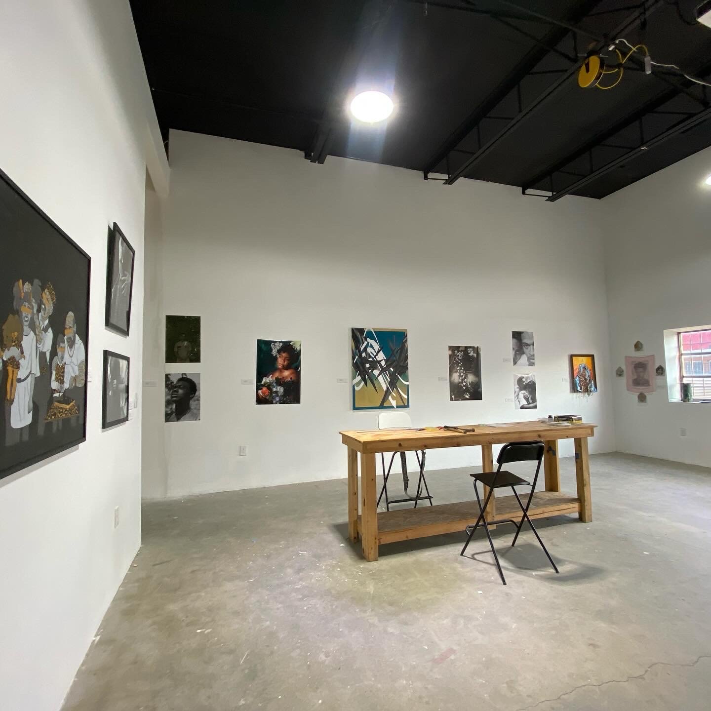 An art gallery with white walls displaying various framed photographs and artwork. A wooden table with a chair and a folding chair in front of it is in the center of the space. There is a window on the right side and ceiling lights illuminating the gallery.