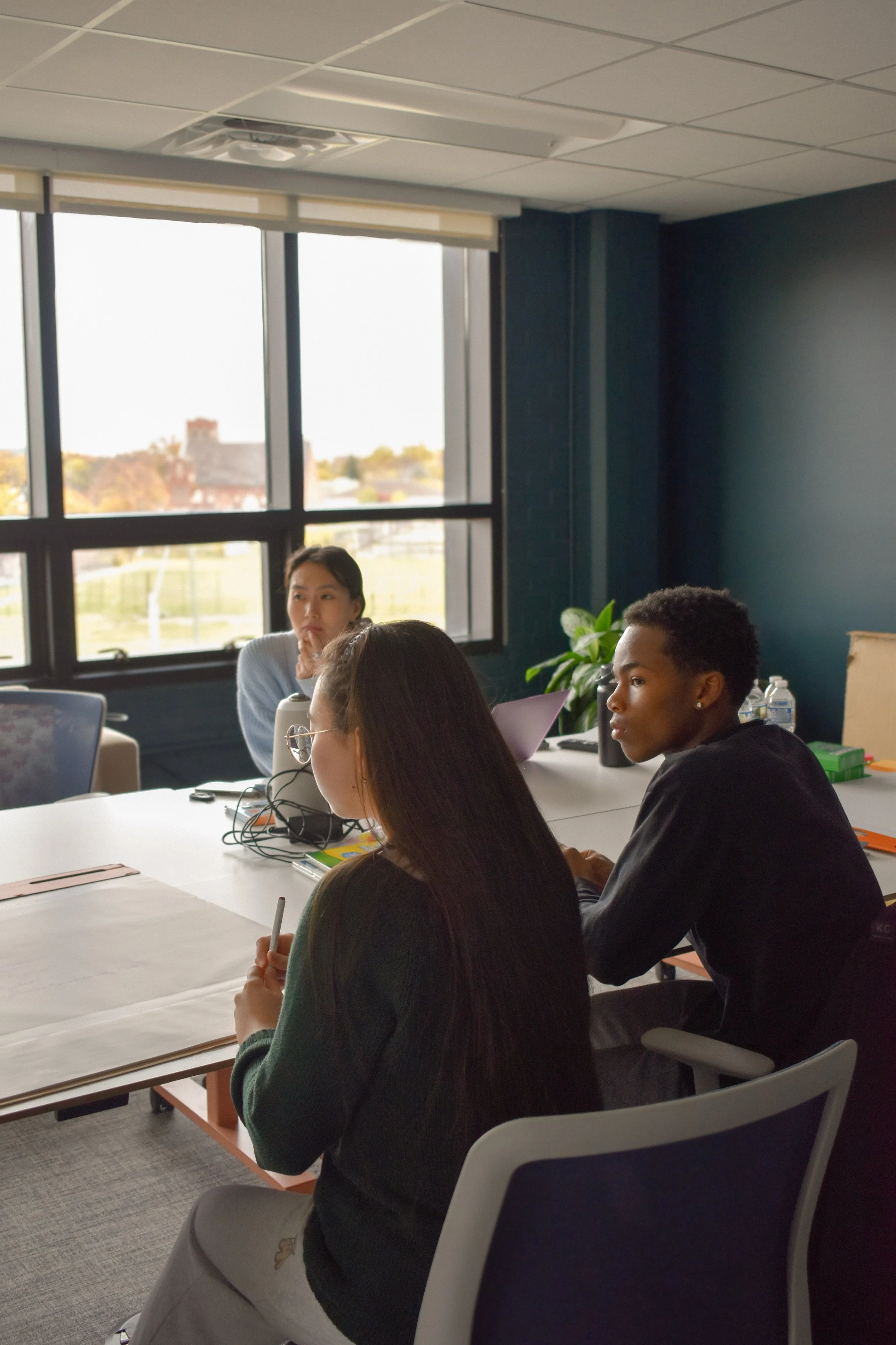 People sitting at a conference table with a large window showing a building and trees outside.