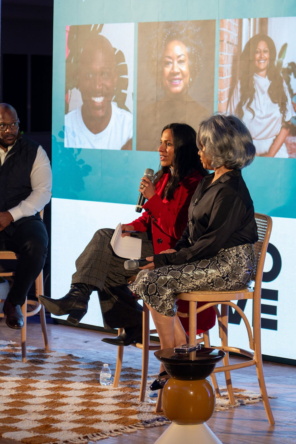 A panel discussion with three women seated on stage, one speaking into a microphone, with a large screen behind them displaying images of three different women.