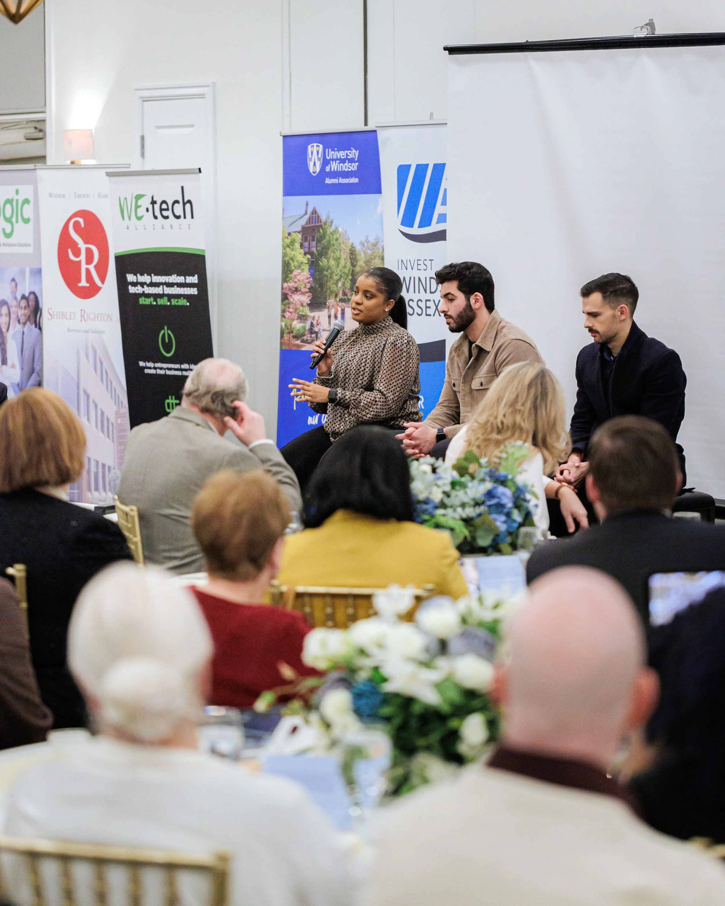 A woman speaking into a microphone during a panel discussion at an event. Three individuals are seated on stage with banners from University of Windsor, WEtech Alliance, and other organizations displayed behind them. Audience members are visible in the foreground.