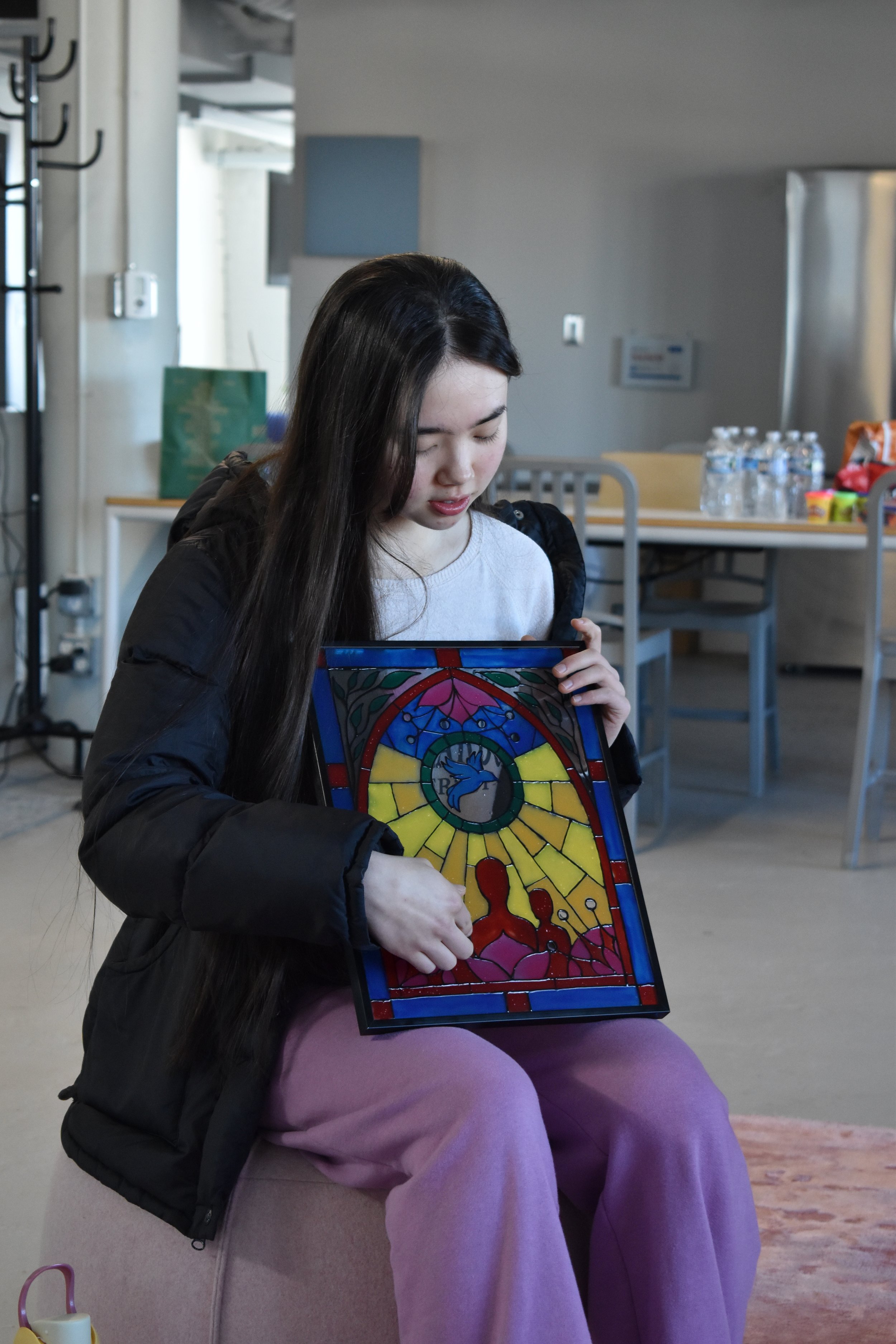 A girl sitting on a person's lap holding a colorful stained glass artwork, with water bottles and snacks on a table in the background.