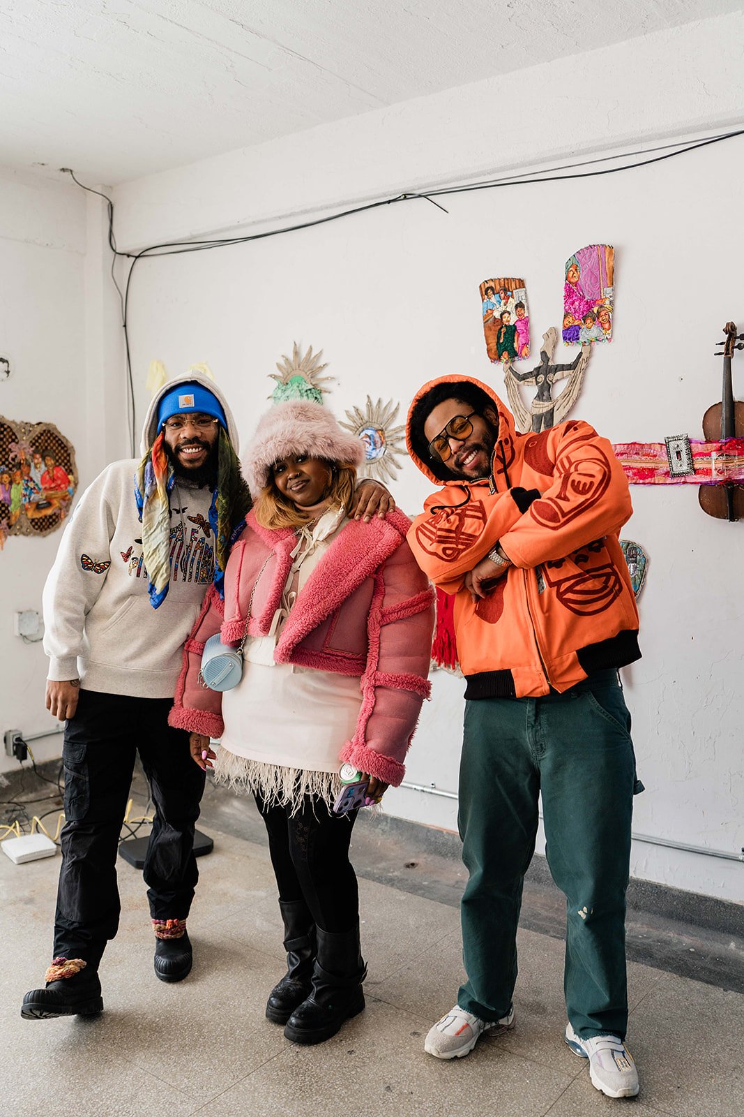 Three people standing together indoors, smiling at the camera, dressed in colorful, winter-style clothing. Behind them on the wall are artistic decorations including a musical instrument and photographs.