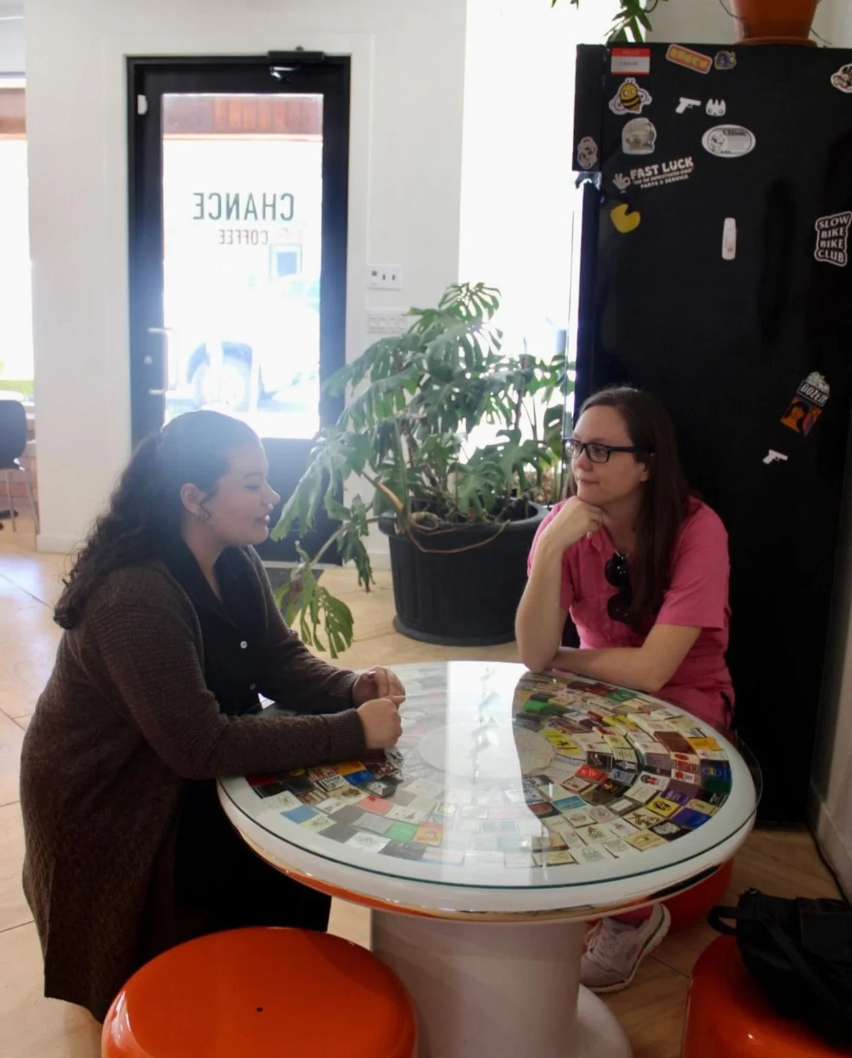 Two women sitting at a round table with a collage of vintage matchbook covers on its surface, engaged in conversation inside a cafe. A large plant and a refrigerator with stickers are in the background, along with a door with the word "CHANGE" on it.