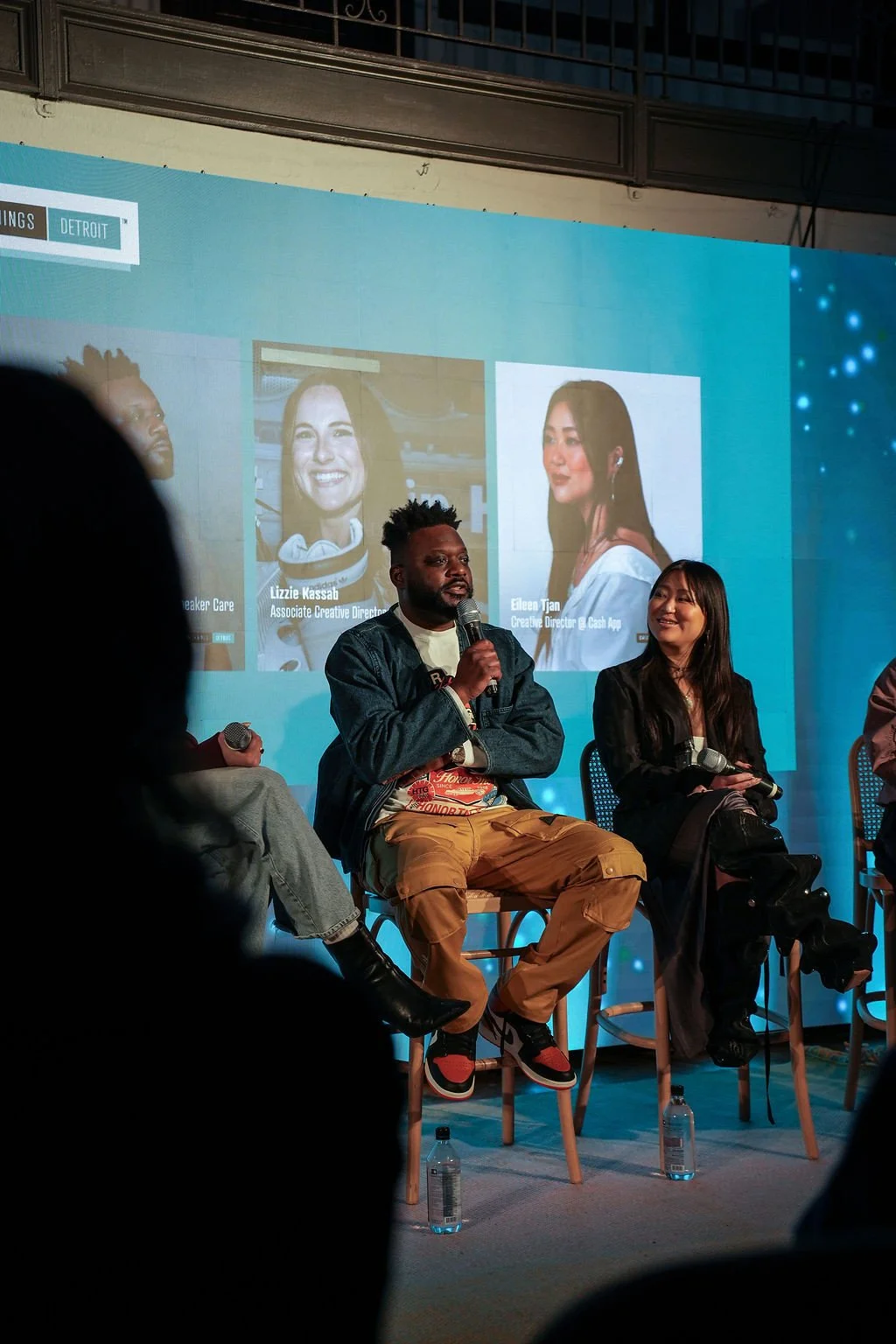 Panel discussion with three people on stage, two women and one man, seated on chairs, with microphones. Large screen behind them displays images and names of two women, Lizzie Kassab and Eileen Tjan, with their titles. The setting appears to be a conference or event in Detroit.