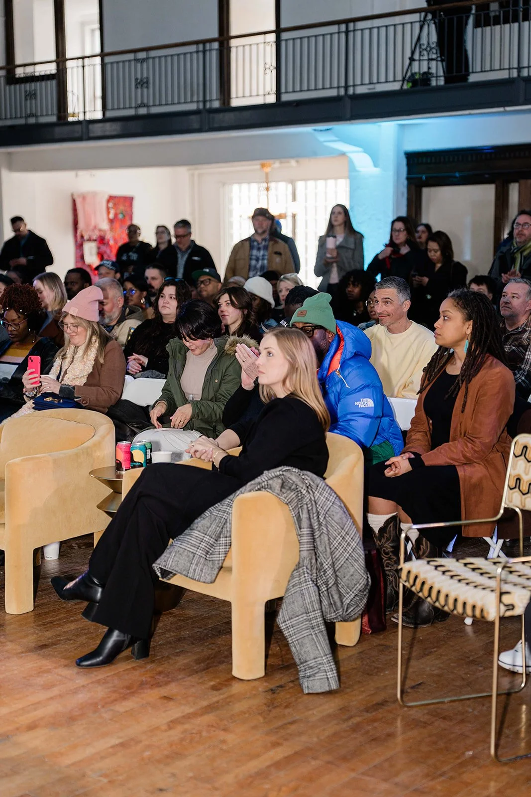 Audience gathered in a spacious, well-lit room, sitting on chairs and sofas, listening attentively to a presentation or discussion, with some standing at the back.