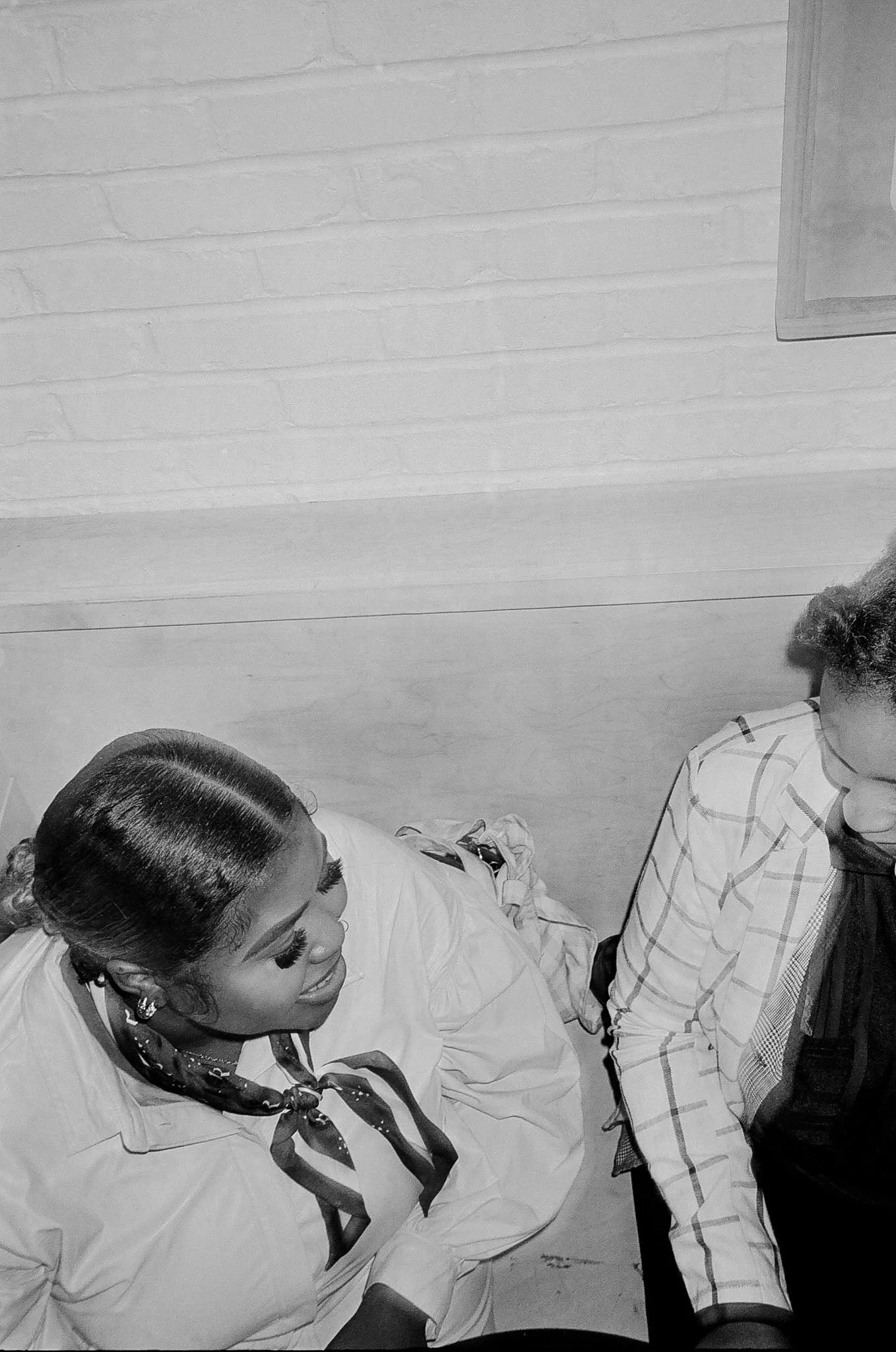 A woman with braided hair, earrings, and a neck scarf smiling and talking to a man wearing a plaid shirt and glasses, in an indoor setting with a brick wall background.