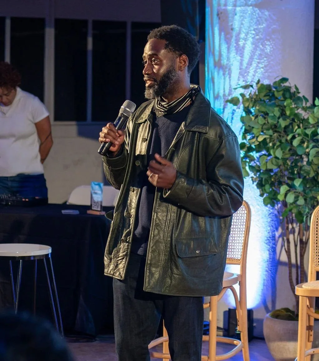 A man with a beard wearing a leather jacket and holding a microphone, speaking at an indoor event with people and chairs visible in the background.