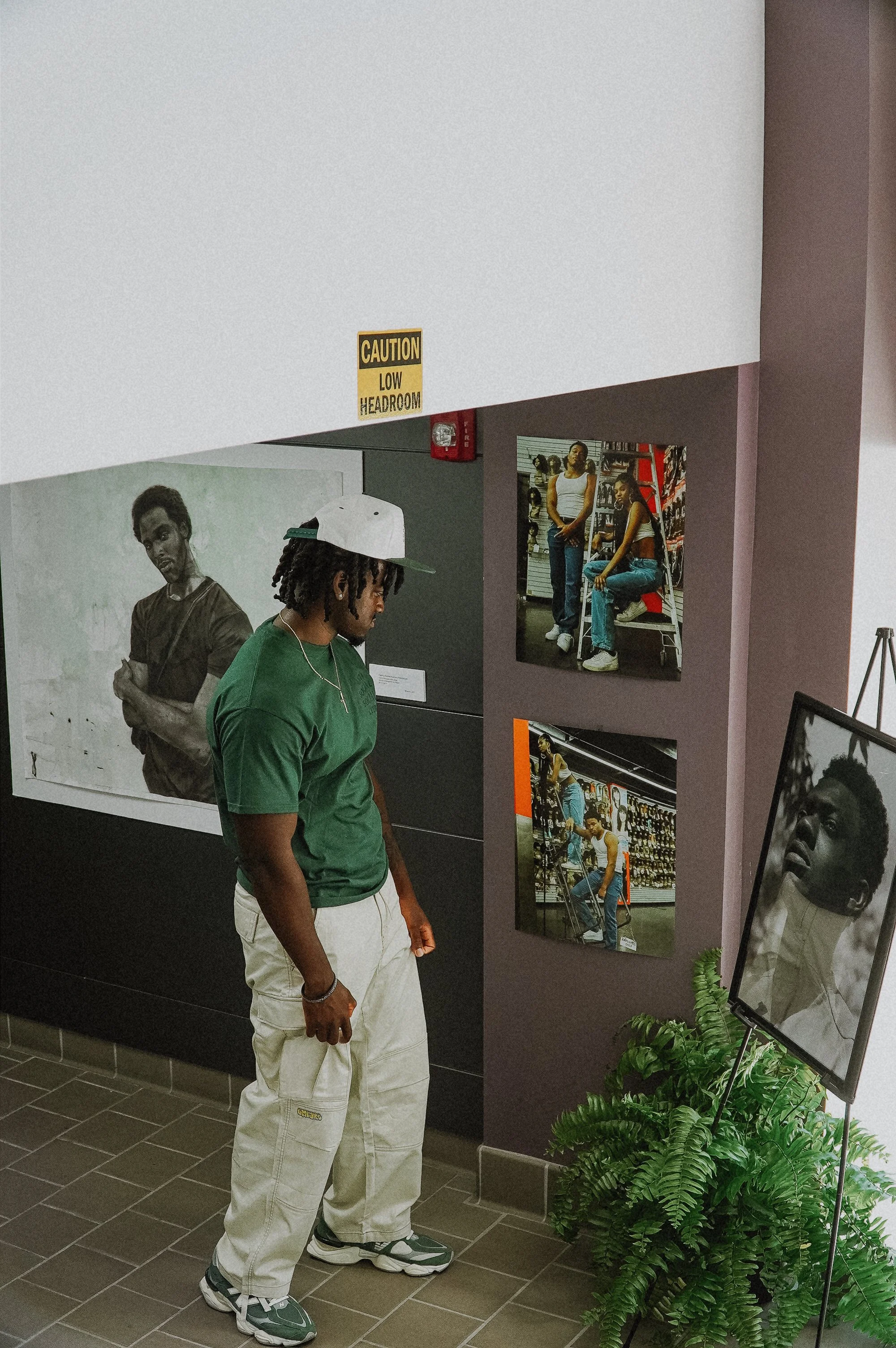 A man with a green t-shirt, white cargo pants, and sneakers standing and looking at photographs on a gray wall at an art or photo exhibition. There are several photos on display, including a black-and-white portrait of a man and a color photo of two women posing inside a store. A plant is on the floor in front of the display, and a mirror or framed photo leans against the wall nearby.
