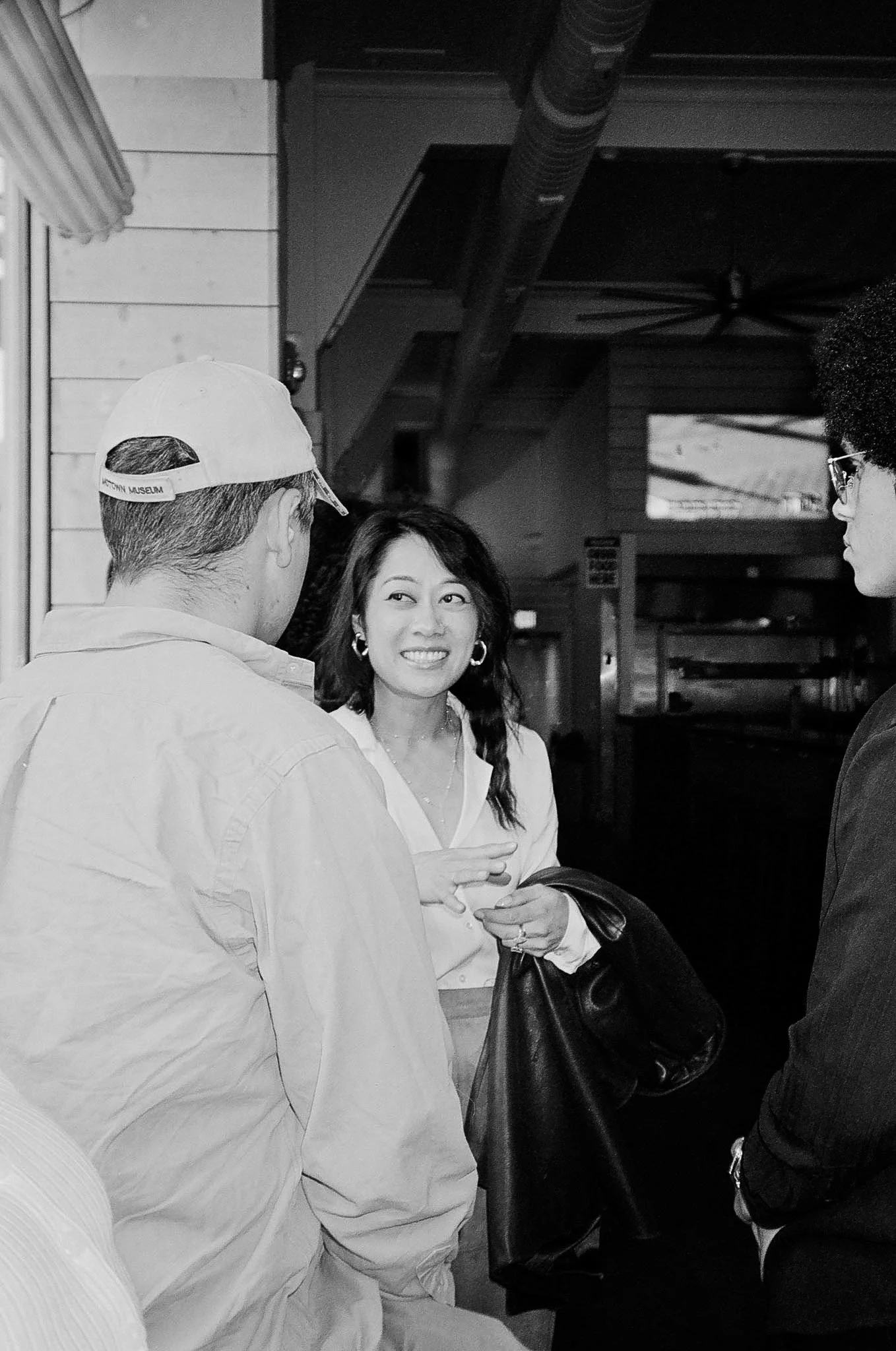 Three people engaged in conversation, two men and one woman, indoors with dark walls, exposed ductwork, and a ceiling fan visible in the background.