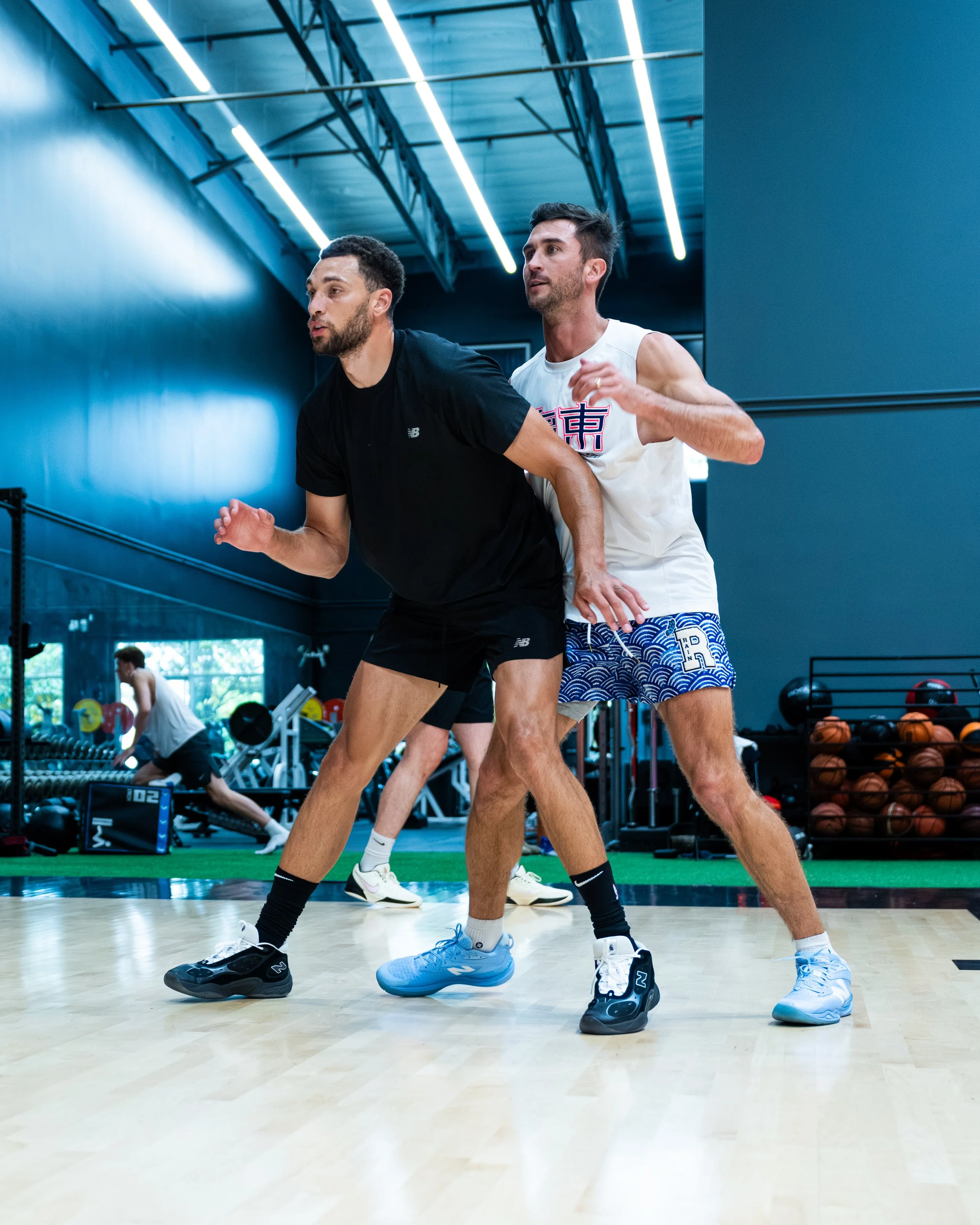 Two men with athletic builds are running in a gym, wearing athletic clothing and sneakers. The gym has high ceilings with exposed metal beams and fluorescent lighting, and sports equipment such as medicine balls, weight racks, and basketballs are visible in the background.