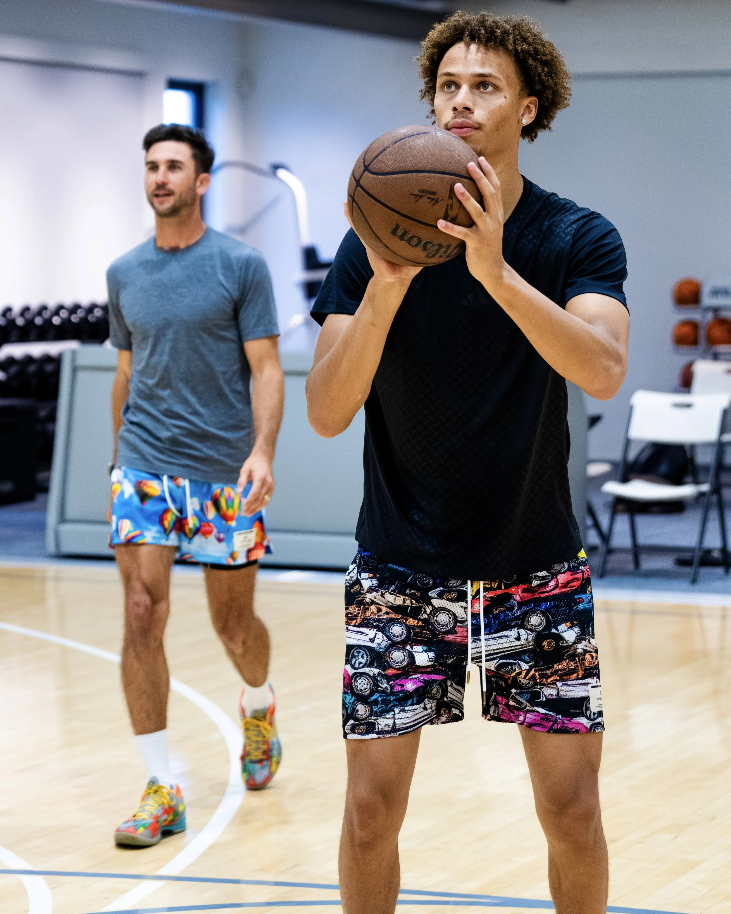 Two men playing basketball in a gymnasium, with one in the foreground holding a basketball and the other in the background running.