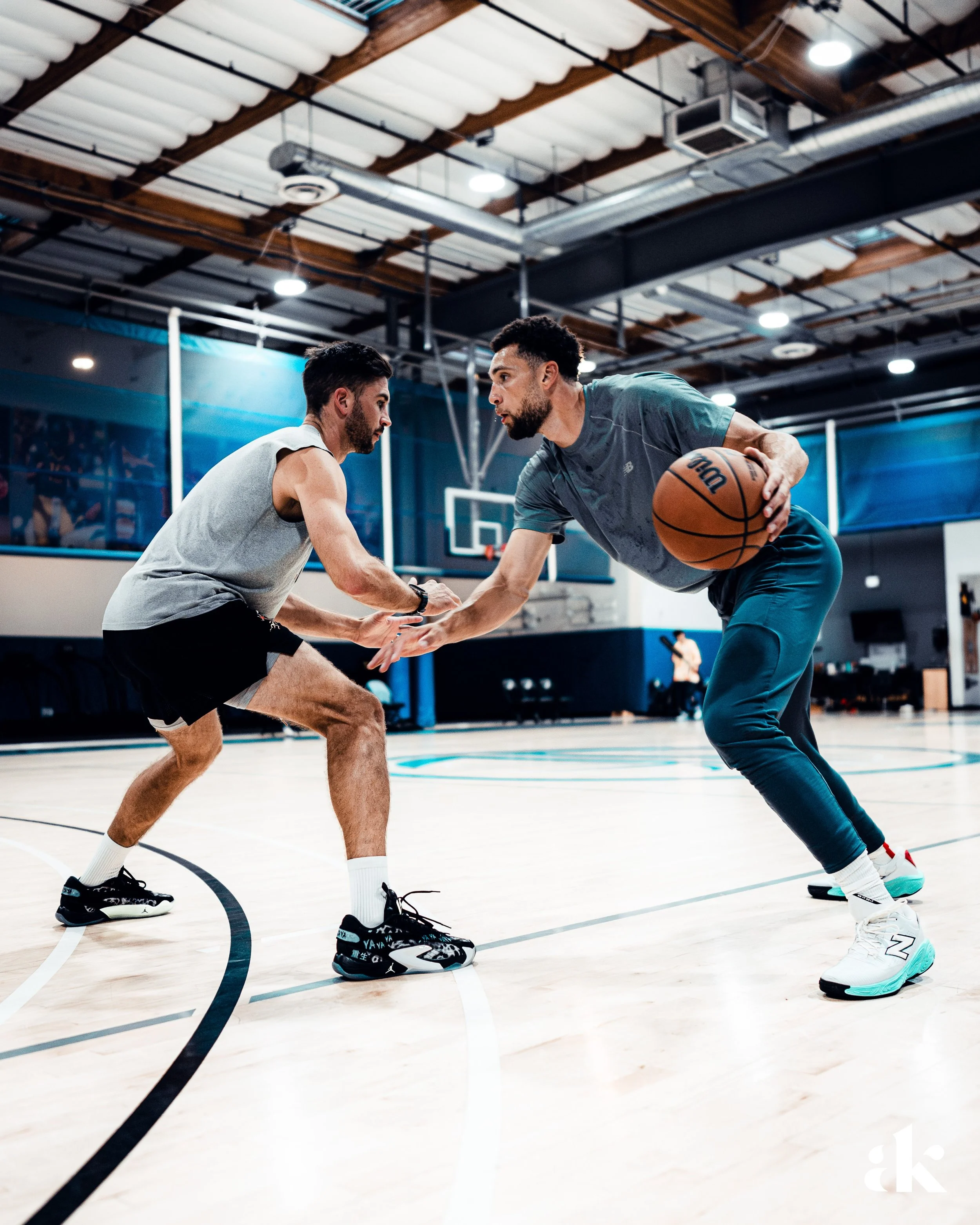 Two men playing basketball on an indoor court, one man dribbling the ball while the other attempts to defend him. The court has a basketball hoop and high ceilings with fluorescent lights.