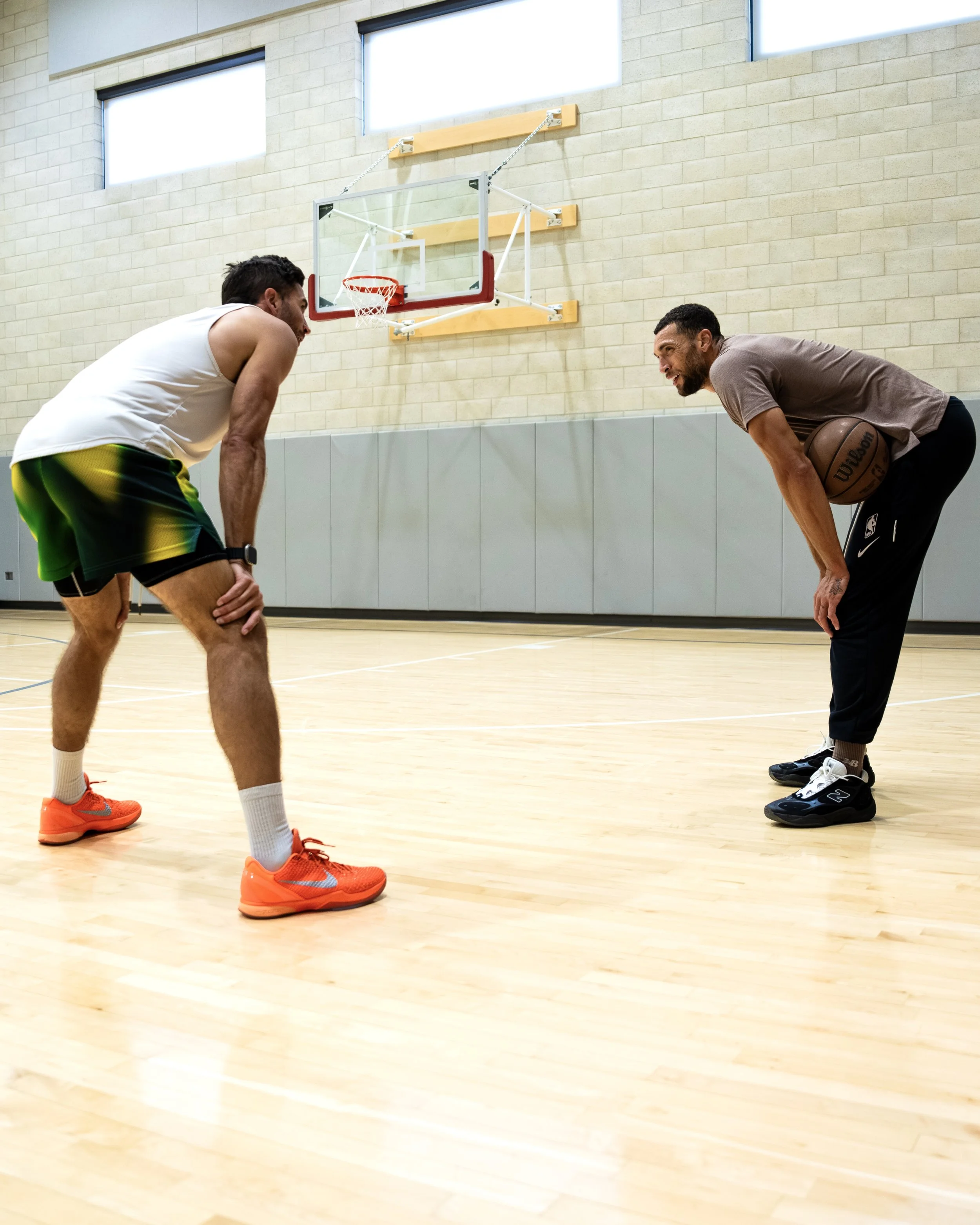 Two men playing one-on-one basketball in an indoor court, both bent forward in defensive stances, facing each other.