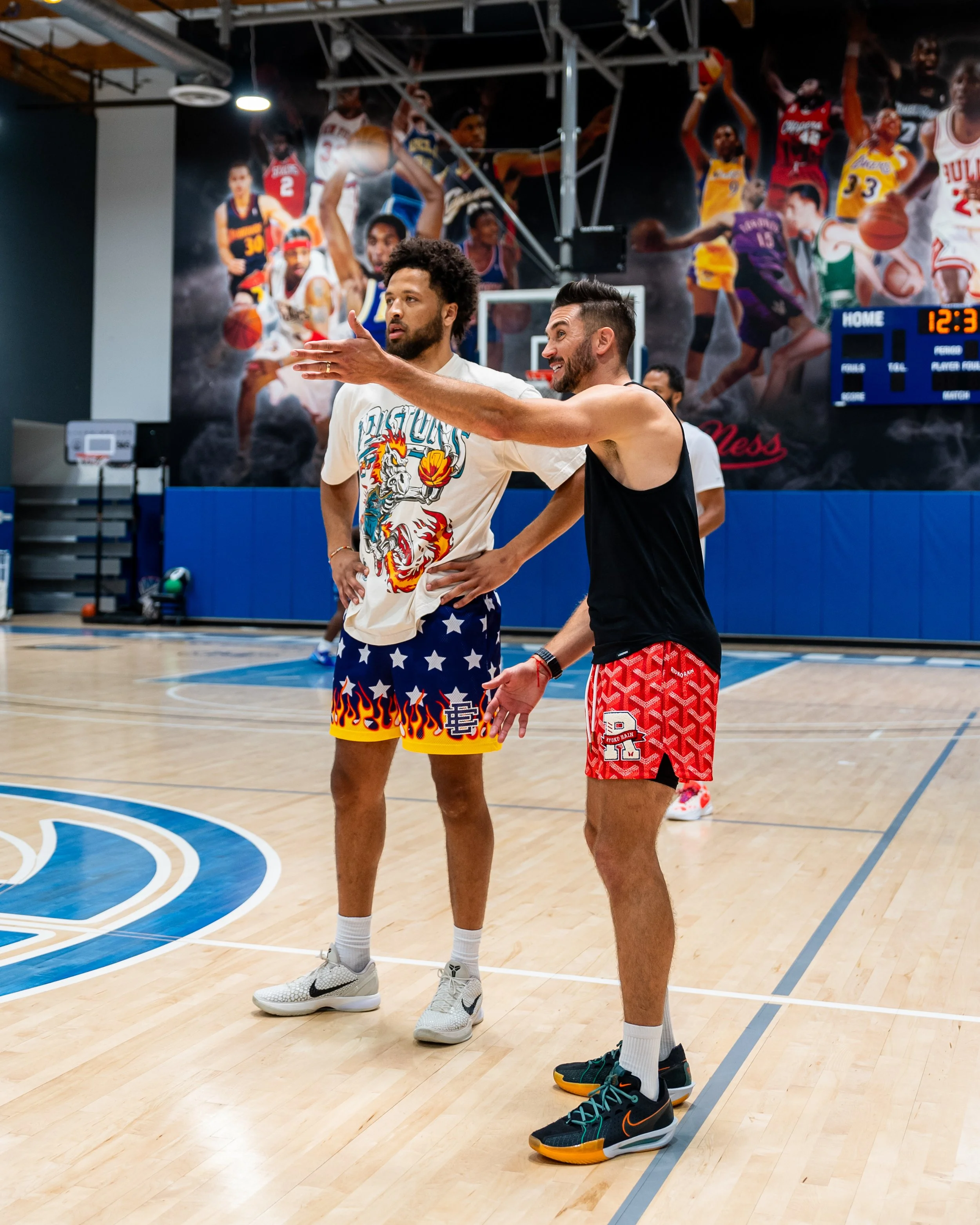 Two basketball players stand on a basketball court, gesturing and talking to each other; a large wall mural featuring various basketball players in action is in the background. The player on the left is wearing a colorful shirt with a dragon and stars design and shorts with flames, while the player on the right wears a black tank top and red patterned shorts. Both are wearing athletic shoes.