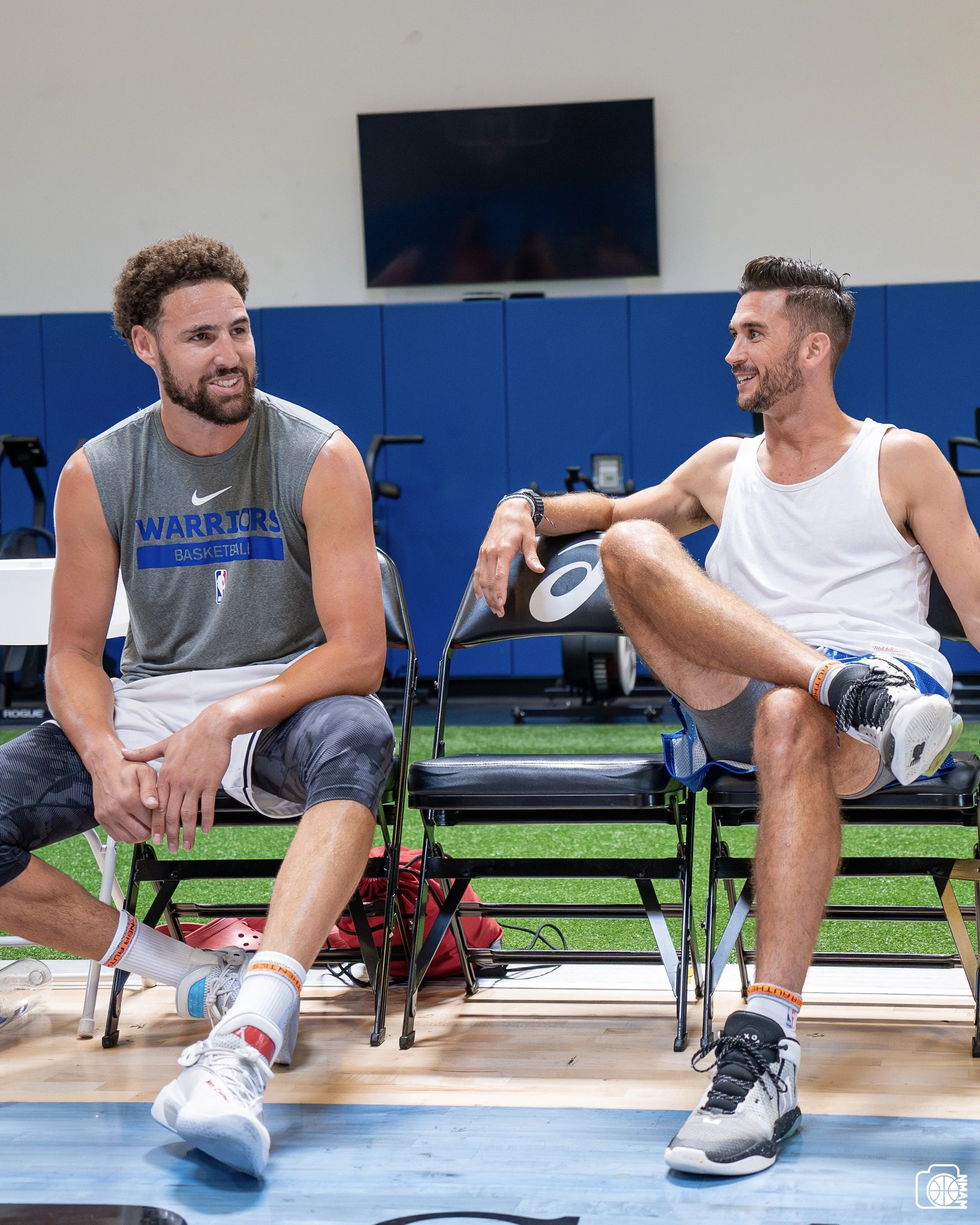 Two men sitting on chairs in an indoor basketball court, smiling and talking to each other. One wears a gray sleeveless shirt and black shorts, and the other wears a white sleeveless shirt and gray shorts. They are wearing athletic shoes and wristbands.