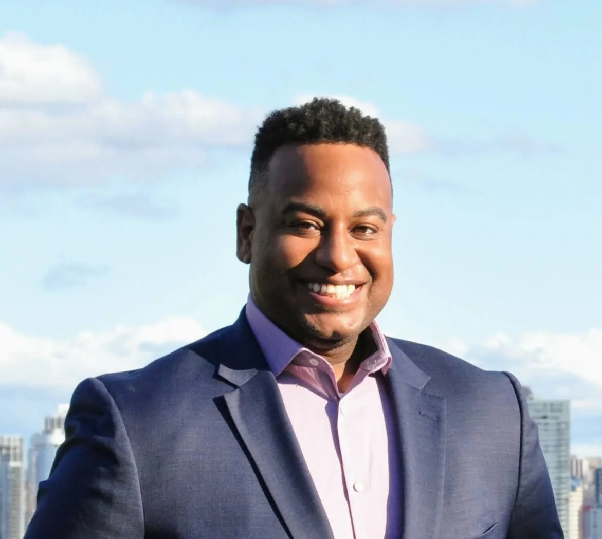 A man in a suit standing outdoors with a city skyline and blue sky with clouds in the background, smiling.