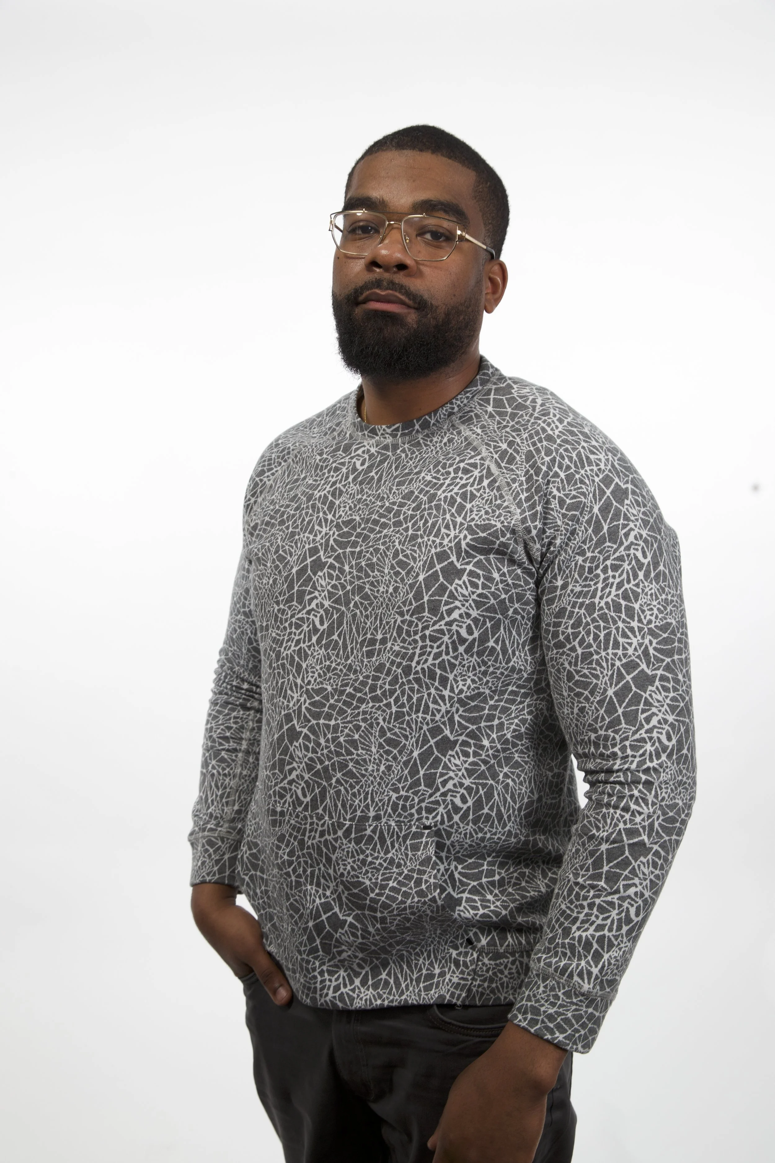 Portrait of a man with glasses, a beard, and dark hair, wearing a white and gray geometric patterned long-sleeve shirt, standing against a plain white background.