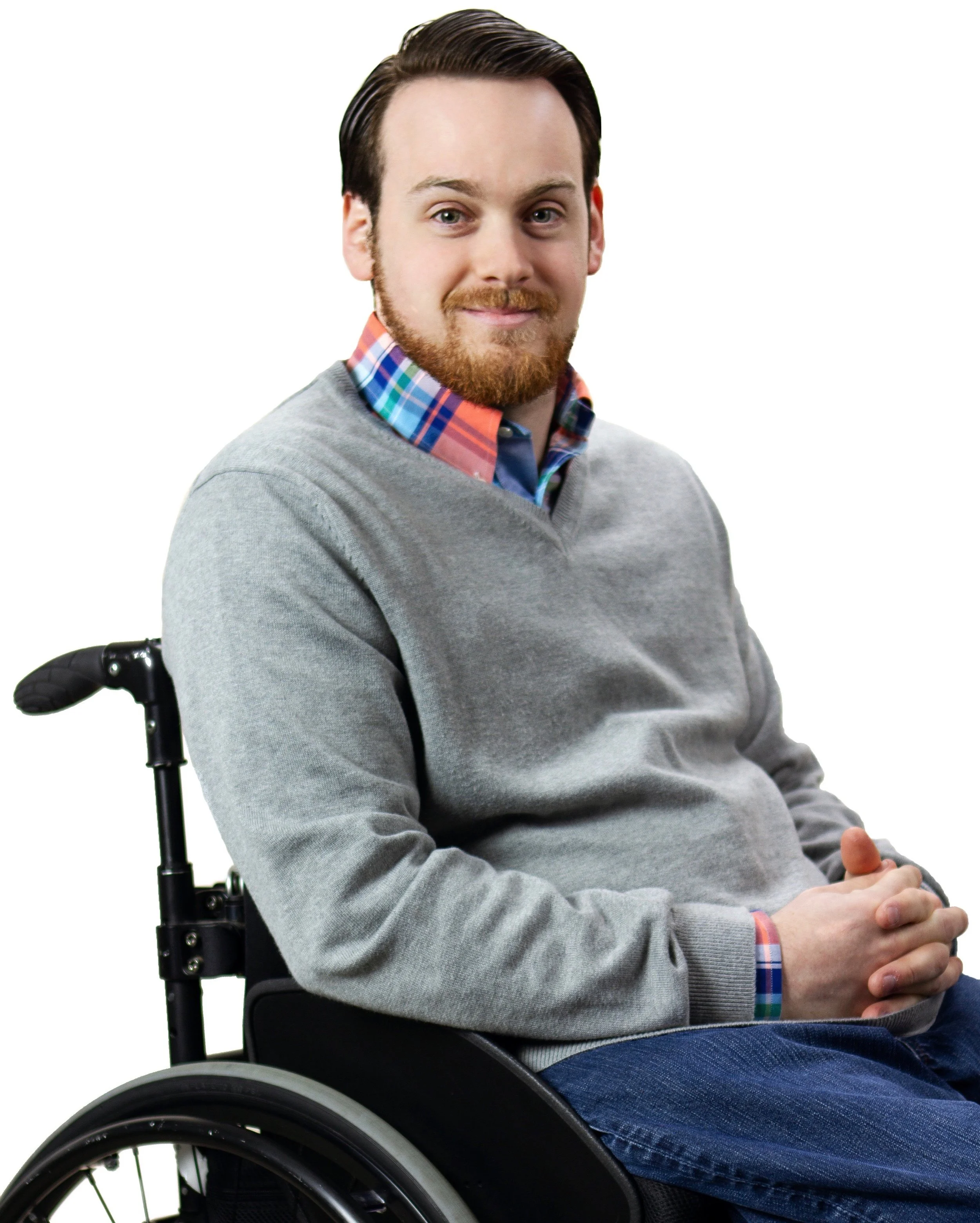 A man with light skin and a beard sitting in a wheelchair, wearing a gray sweater over a colorful plaid shirt, smiling softly.