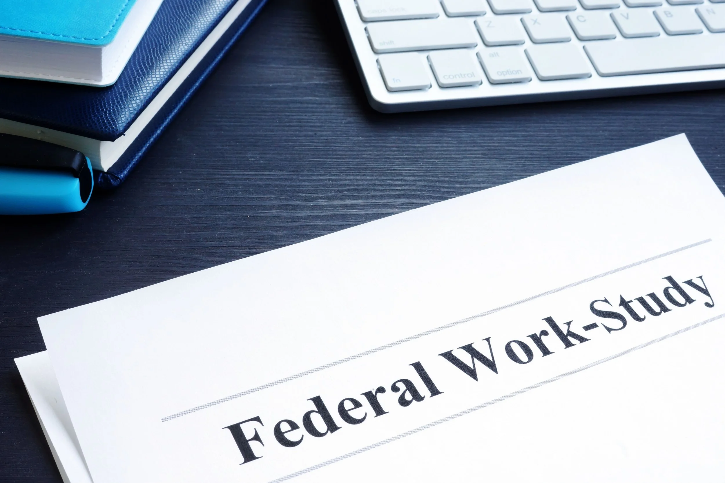 Close-up of a federal tax work-study document on a dark wooden desk, accompanied by a white computer keyboard, blue notebooks, and a blue pen.