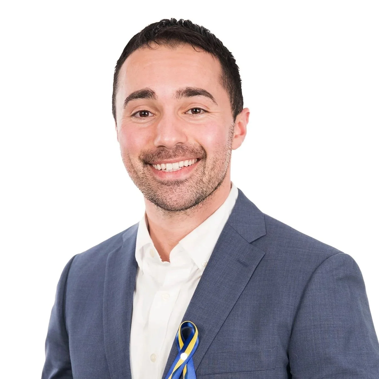 Headshot of a man smiling, wearing a blue suit and white shirt, with a ribbon in the colors black, yellow, and blue.