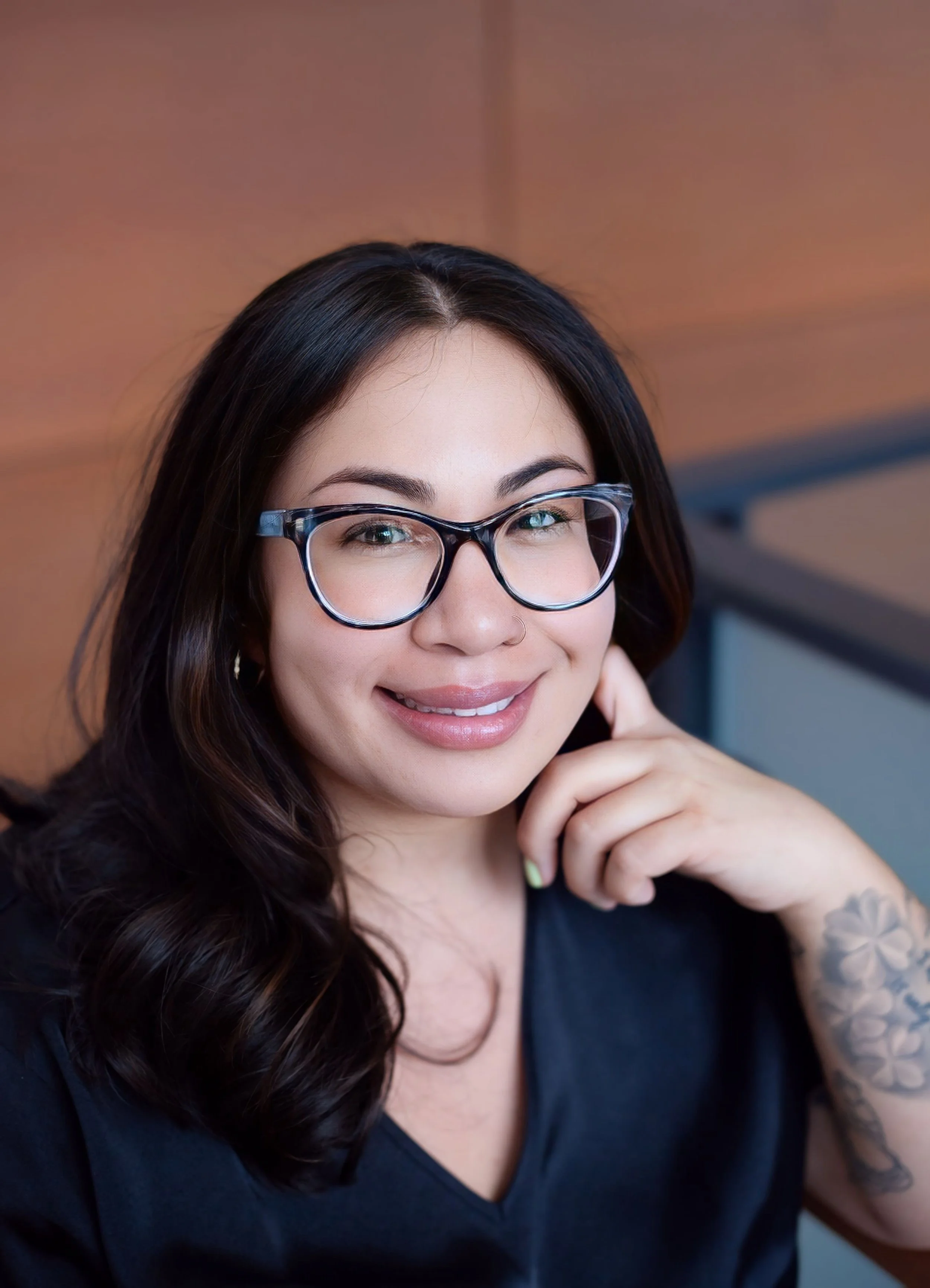 Close-up of a smiling young woman with long dark hair, wearing glasses, and a tattoo on her arm, touching her face.