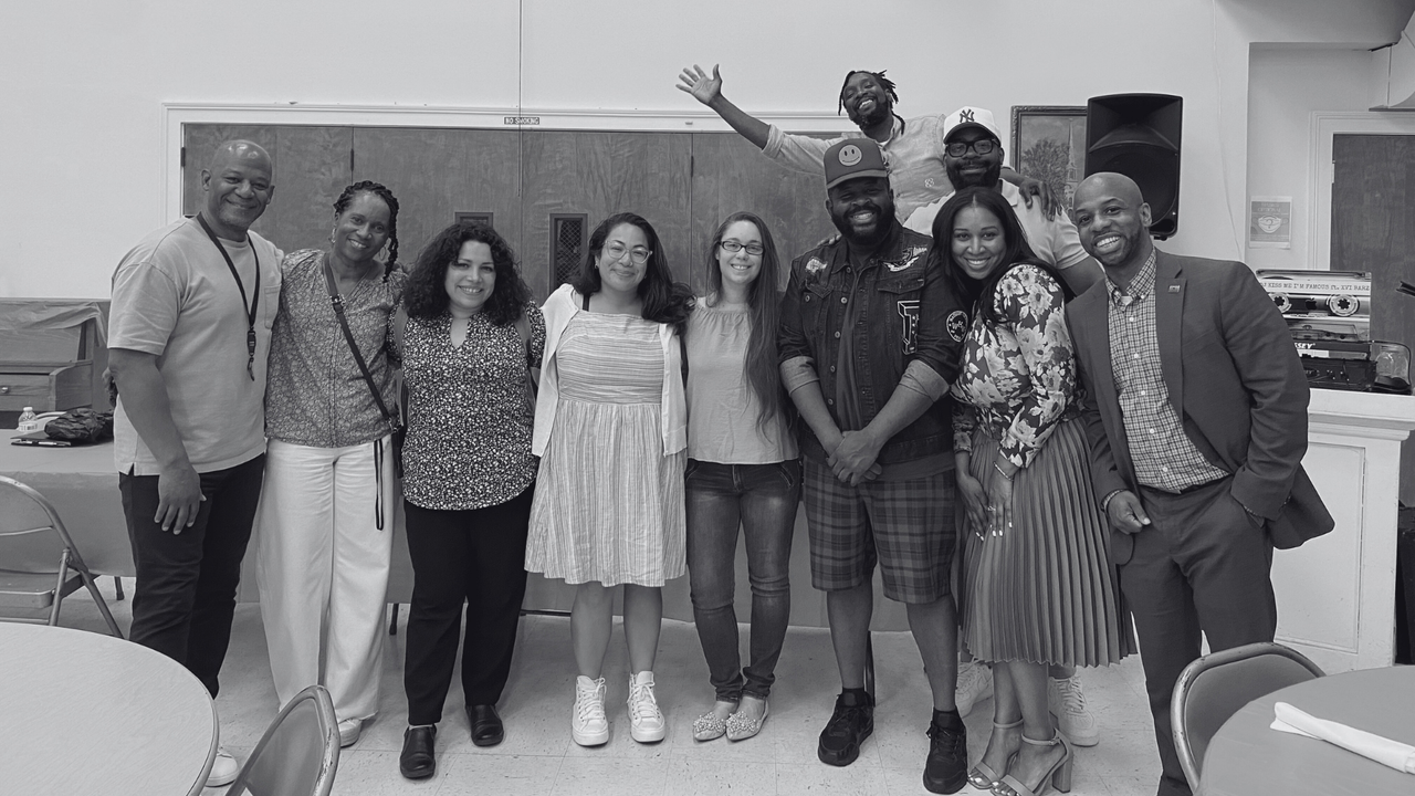 Group of 10 diverse people smiling and posing together in a room, some with arms around each other, some making gestures, with chairs and tables in the foreground.