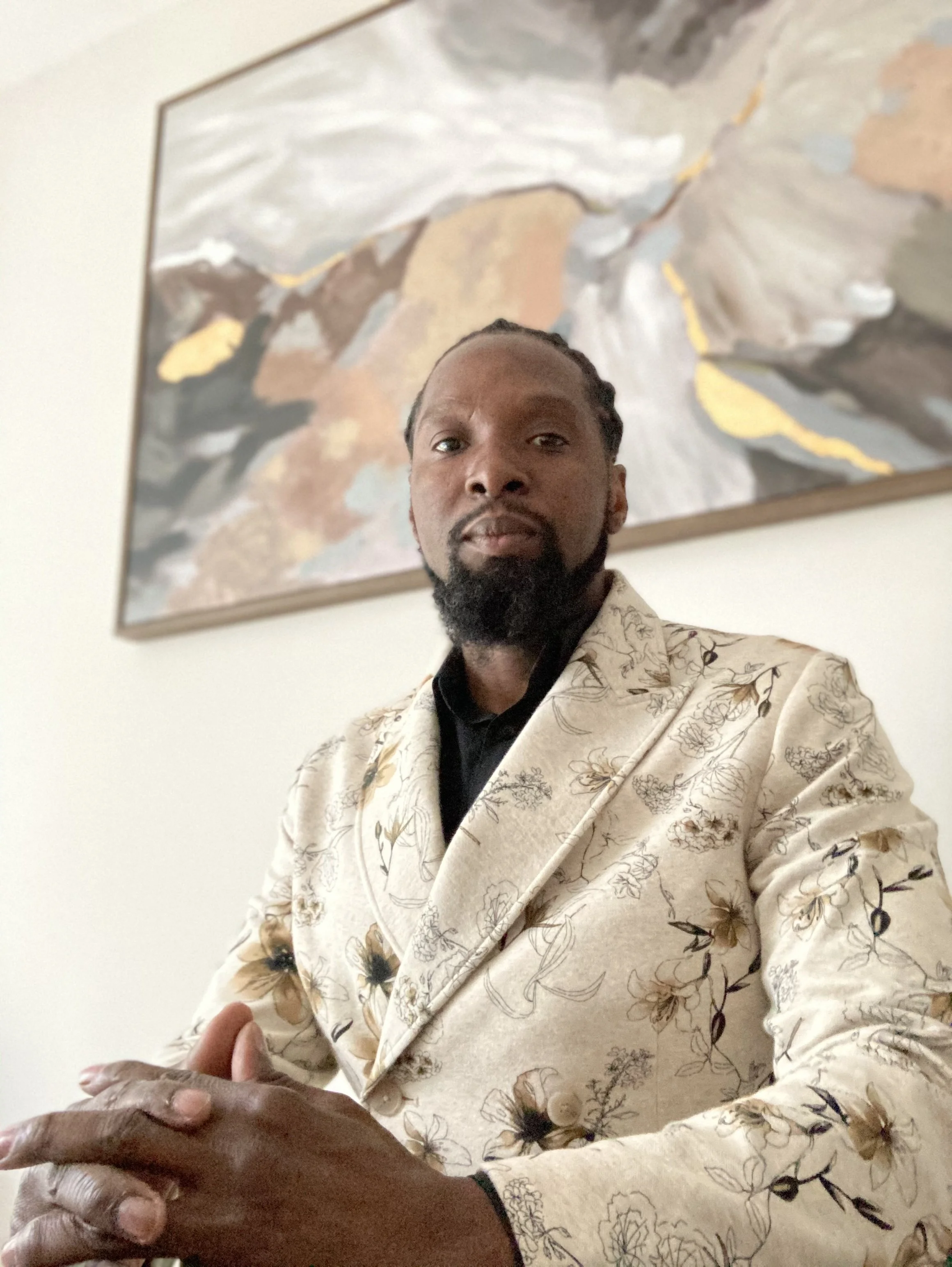 A man with dark skin, a beard, and braided hair wearing a cream-colored blazer with a floral pattern, standing in front of an abstract painting with earth tones and white accents, looking at the camera.