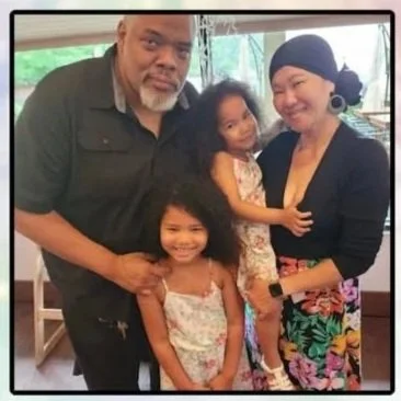 A family of four smiling for a photo indoors. The man has a beard and mustache, the woman and two young girls are happy and posing.