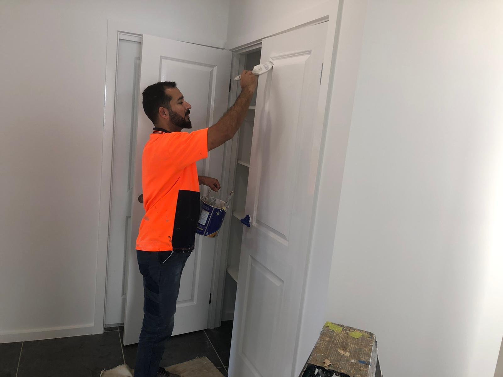 A man wearing an orange shirt is painting a white door with a brush. He holds a paint can in his left hand and is standing on a dark tiled floor, with a white wall and door frame in the background.