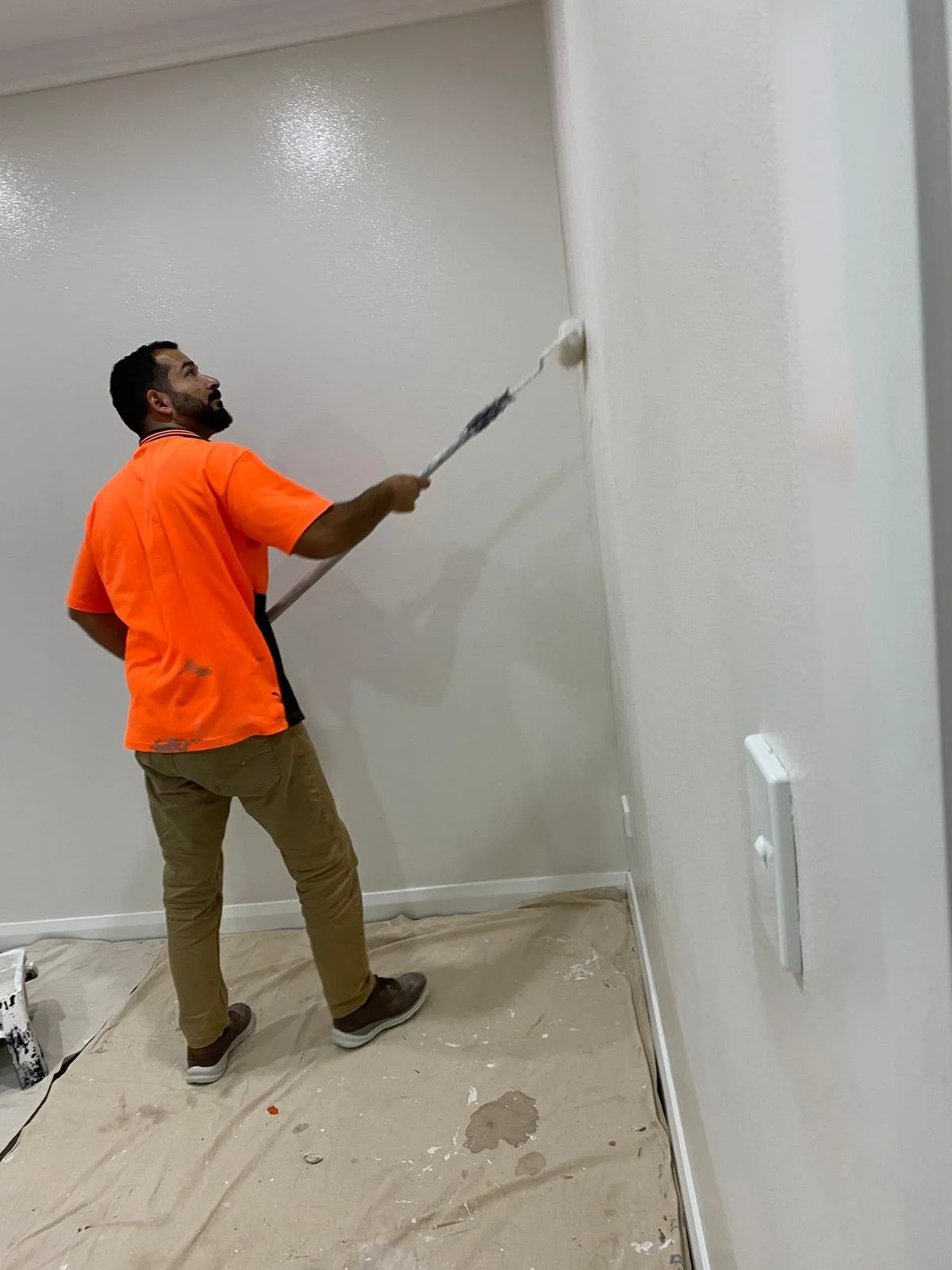 A man wearing an orange shirt and khaki pants is using a paint roller to paint a white wall in a room. There is a plastic sheet on the floor to protect it from paint spills.