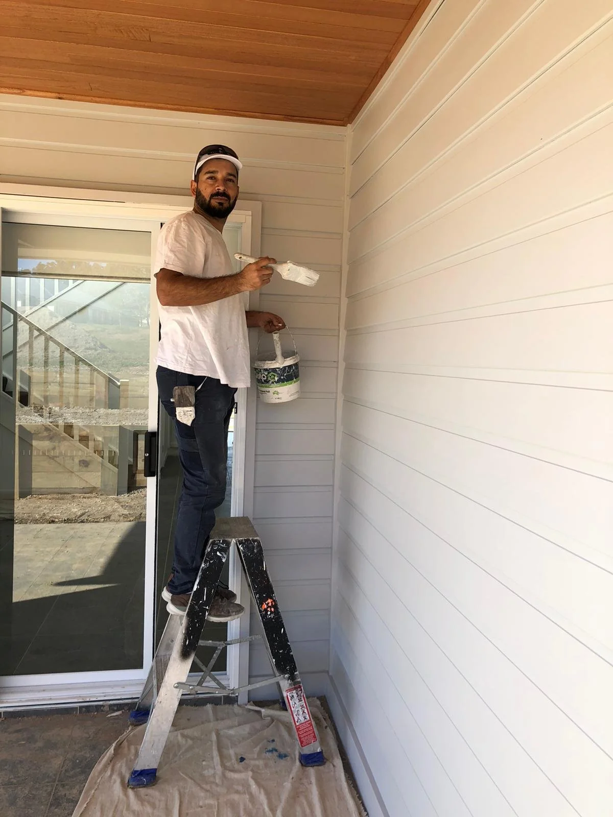 Man painting the exterior wall of a house while standing on a step ladder.
