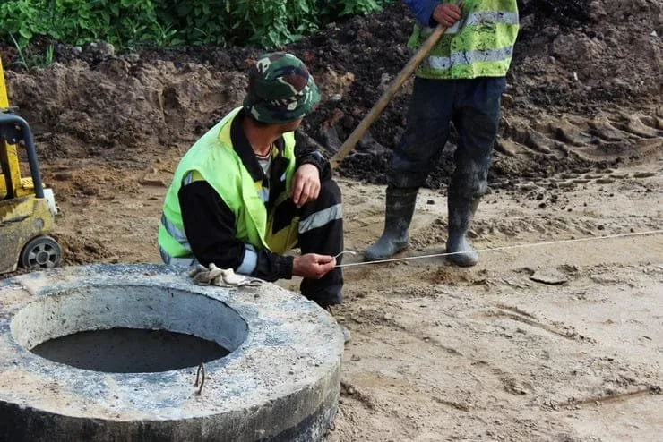 Two construction workers in high-visibility safety vests and boots working outdoors. One is crouched down, holding a measuring tape, while the other stands nearby with a shovel, on a dirt site with greenery in the background.