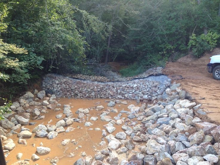 A small creek with rocks used as a culvert for water flow, surrounded by greenery and a dirt road with a parked vehicle on the right.